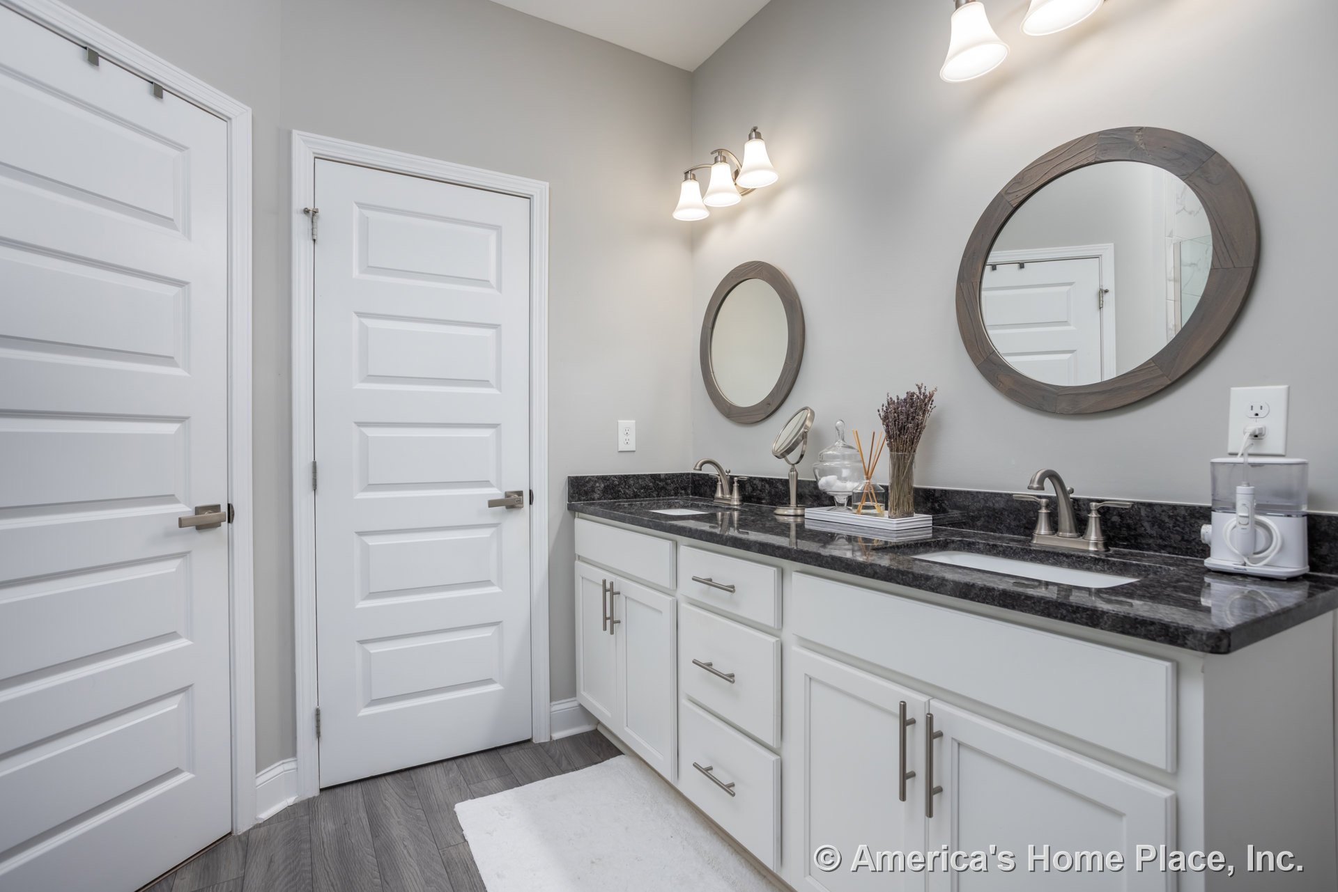 Bathroom with white shaker cabinets, quartz countertop, two wall-mounted mirrors, silver faucet, white door with silver handle, three-light vanity fixture above mirrors, two-light