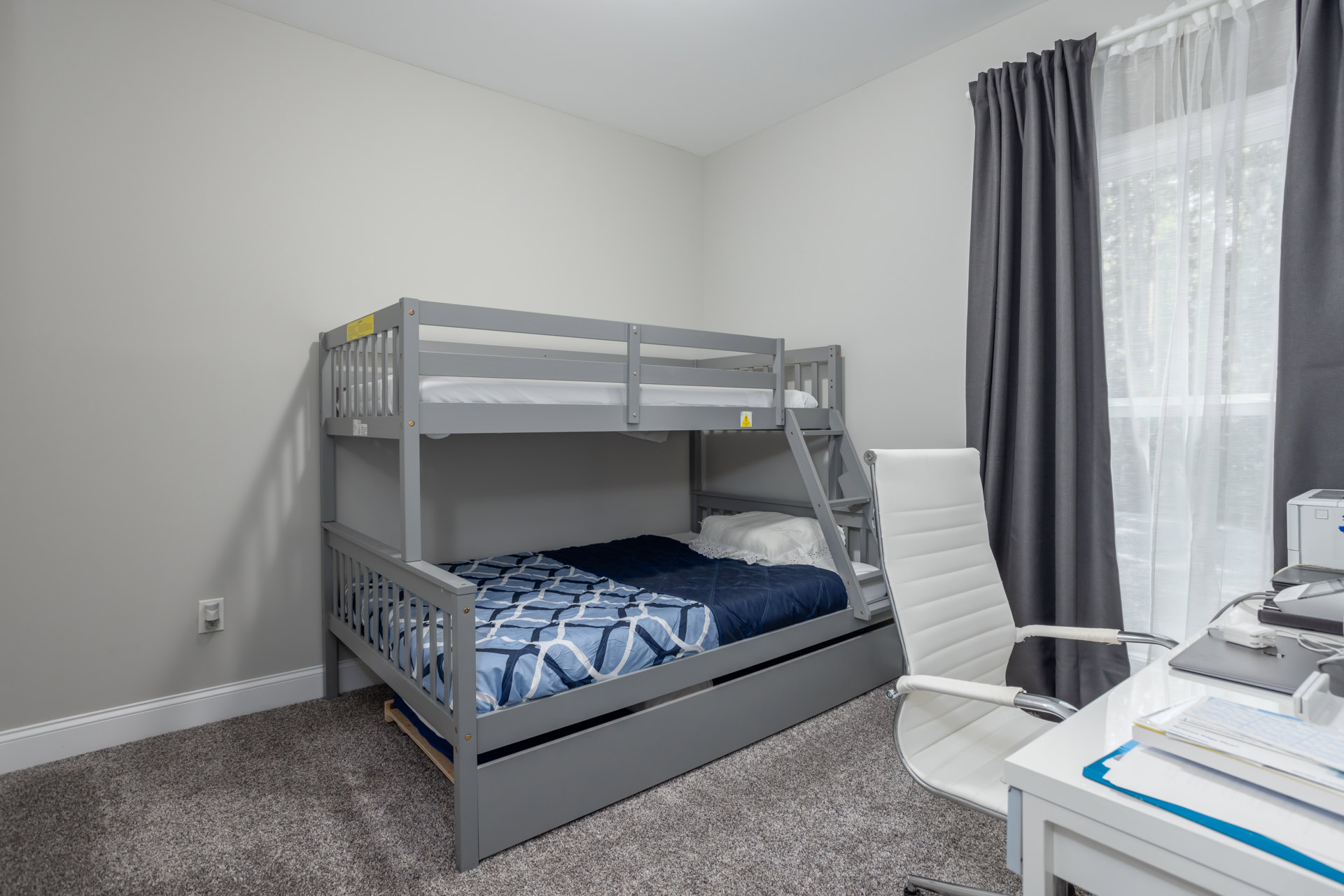 Wooden bunk bed with blue blanket and white pillow beside window with sheer grey curtains, white chair, and light-colored flooring