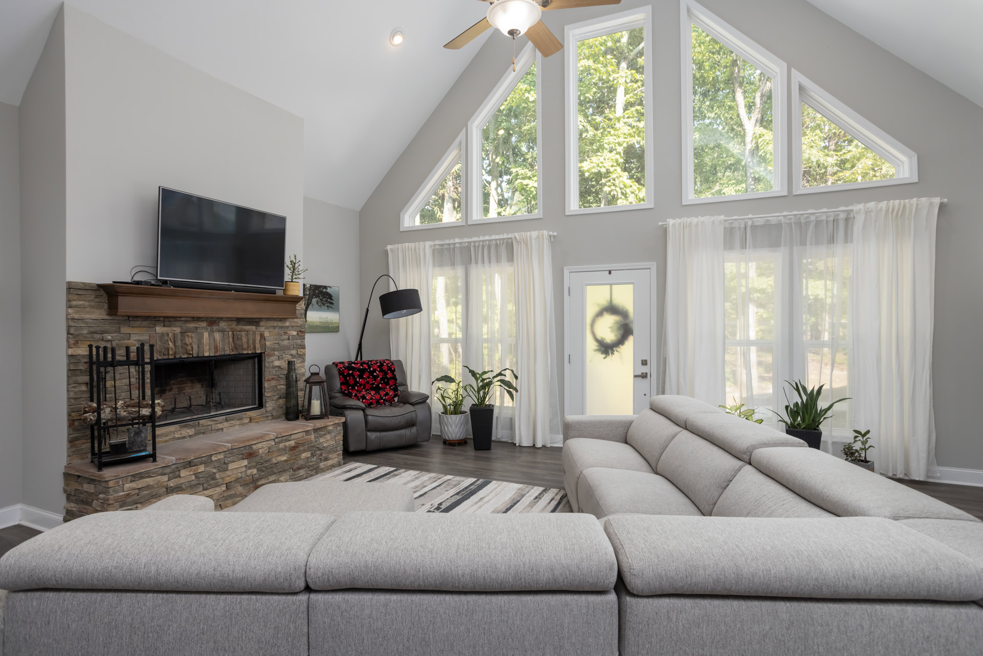 Living room with a stone fireplace, large sectional couch, black television on a table, log rack, and chair draped with a blanket near windows.
