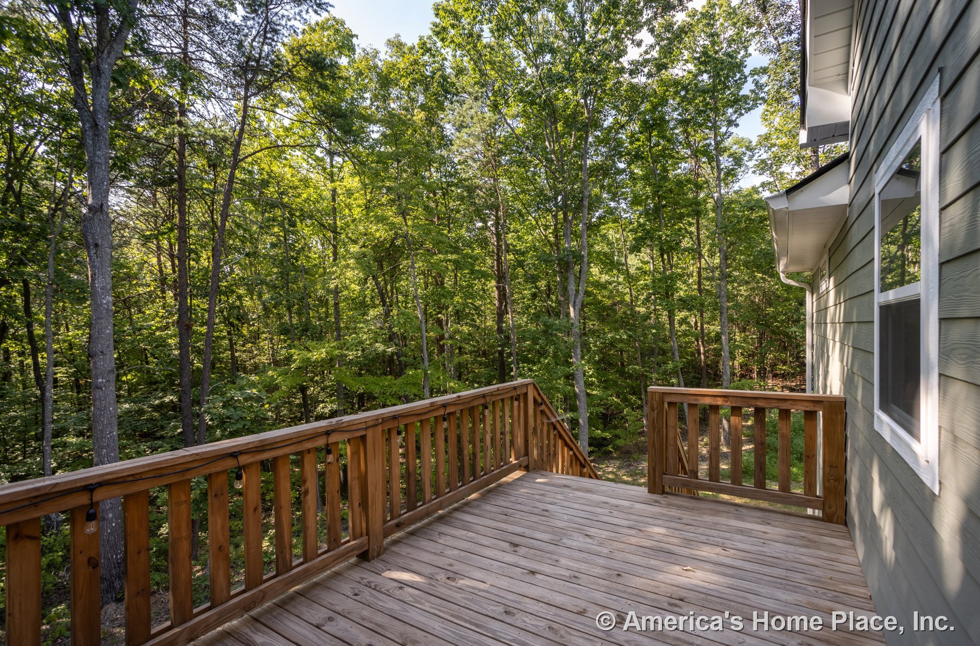 Wooden deck with railing, string lights, and large windows, surrounded by mature trees in the background