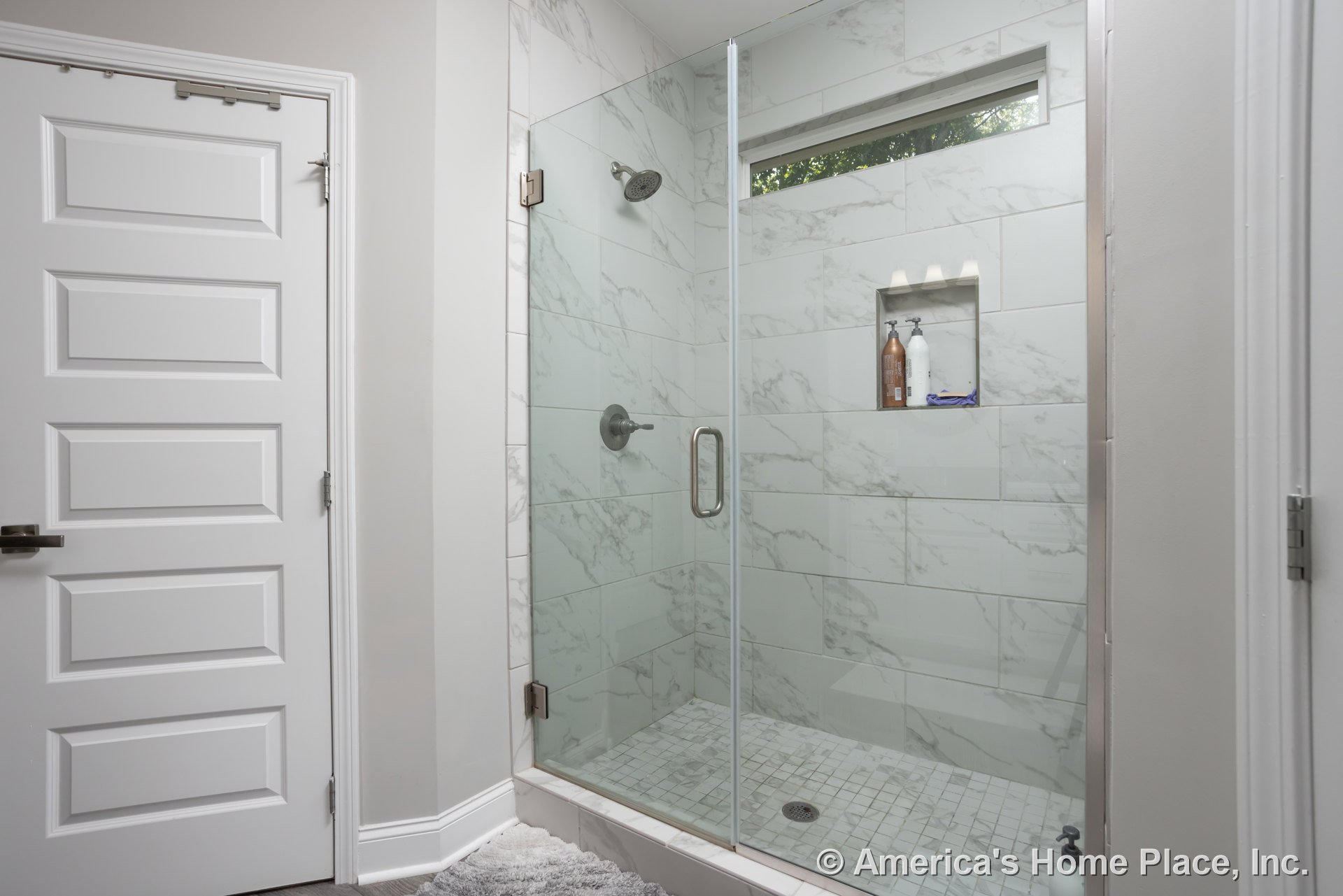 Glass shower enclosure with tiled floor, chrome shower head mounted on wall, white door with metal handle, close-up of modern faucet, visible shower drain