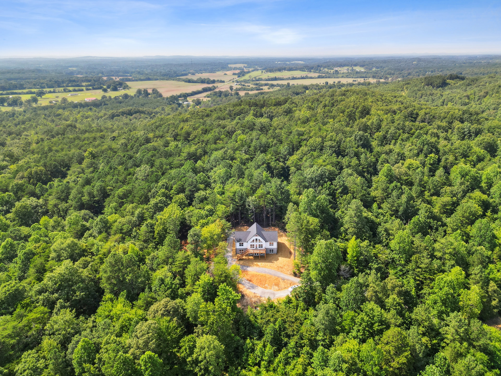 Two-story house with wooden deck and covered porch, nestled among mature trees, blue sky with scattered clouds overhead, landscaped walkway leading to entrance