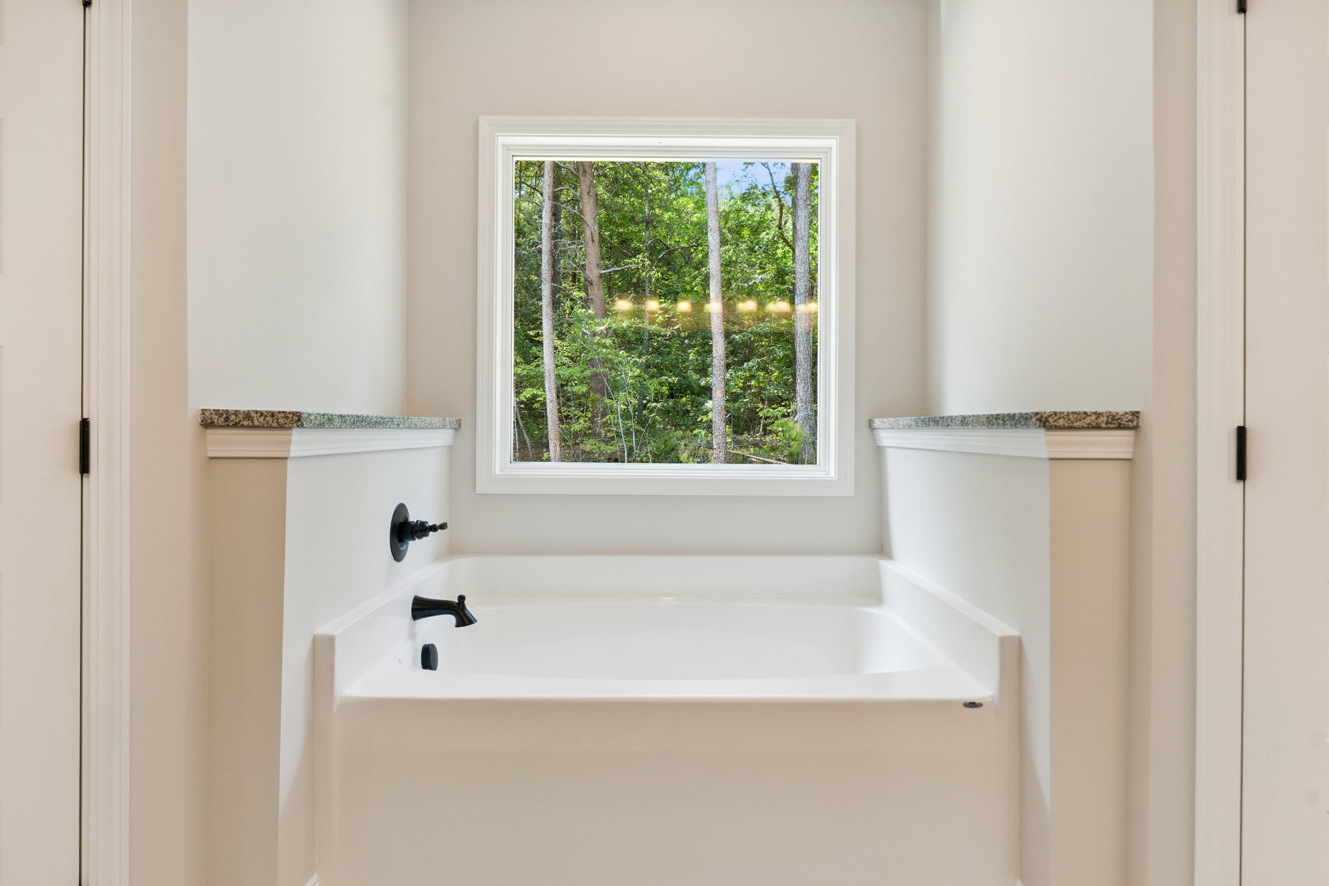 Freestanding white bathtub with black faucet beneath a large window overlooking trees, surrounded by white tile walls in a modern bathroom.