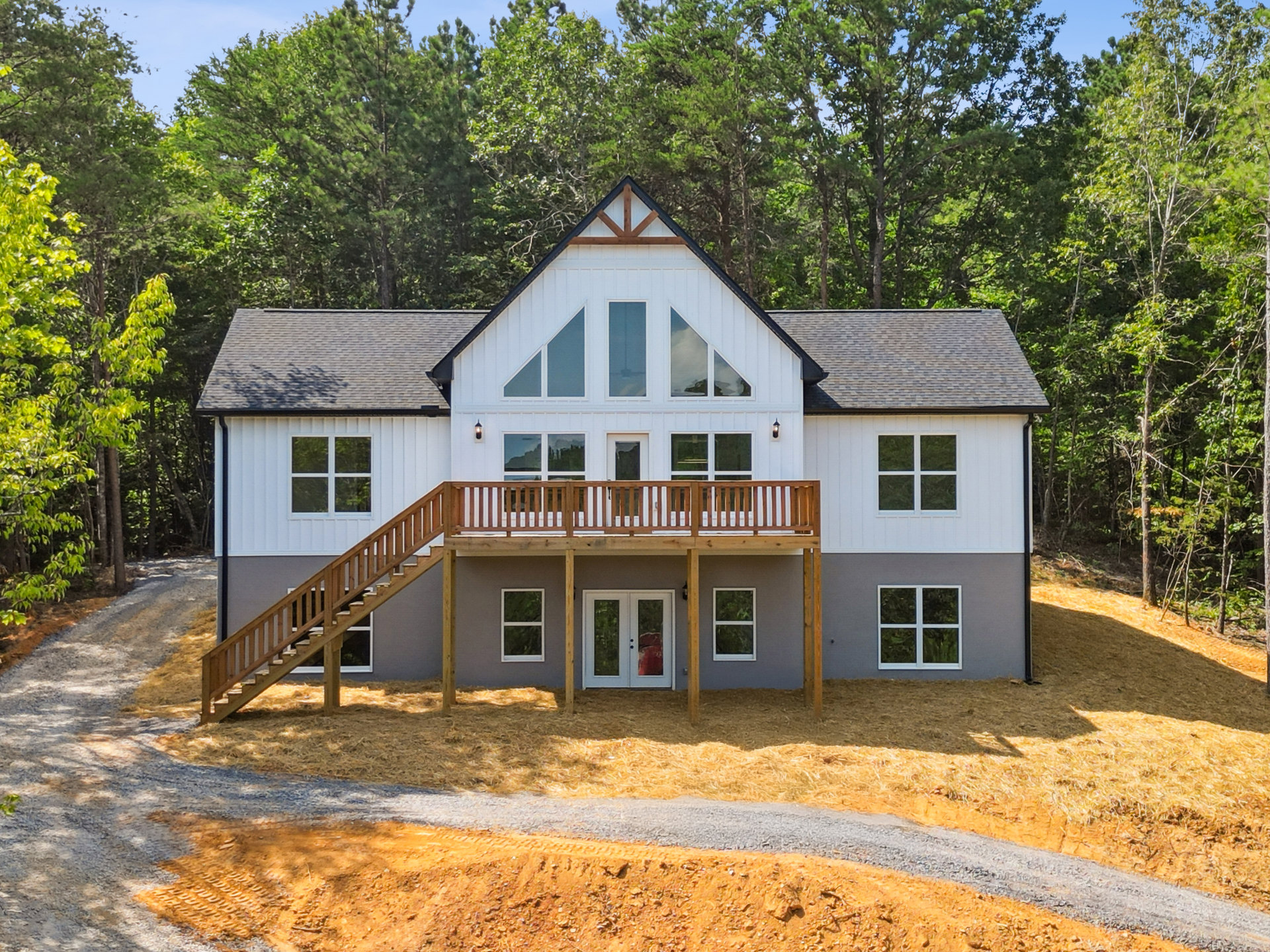 White house with triangular roof, glass-paneled double doors, elevated wooden deck with stairs, white-framed windows, surrounded by trees.