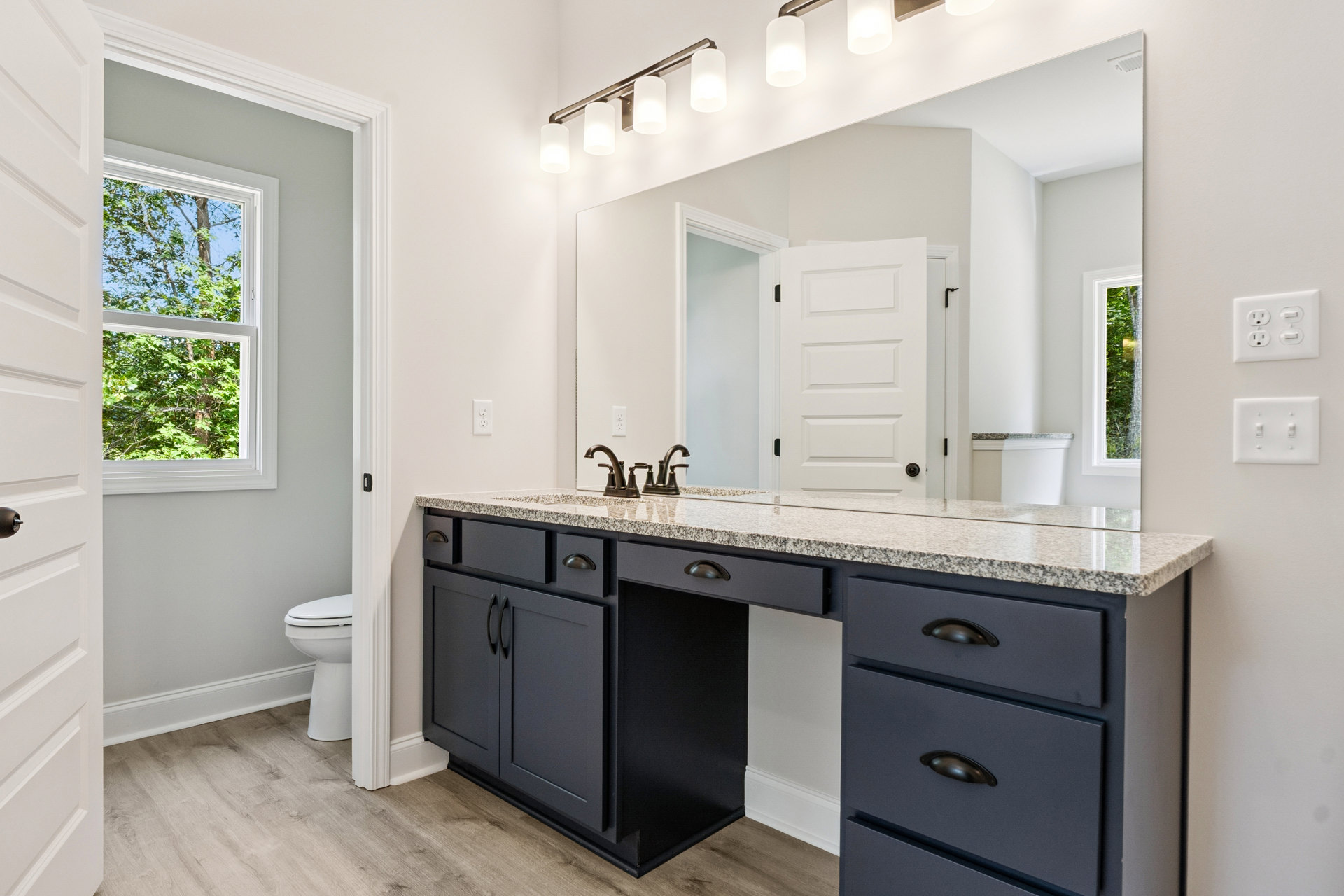 Bathroom with marble countertop, white toilet, wood cabinetry, tile flooring, window showing trees, electrical outlet and light switch on wall