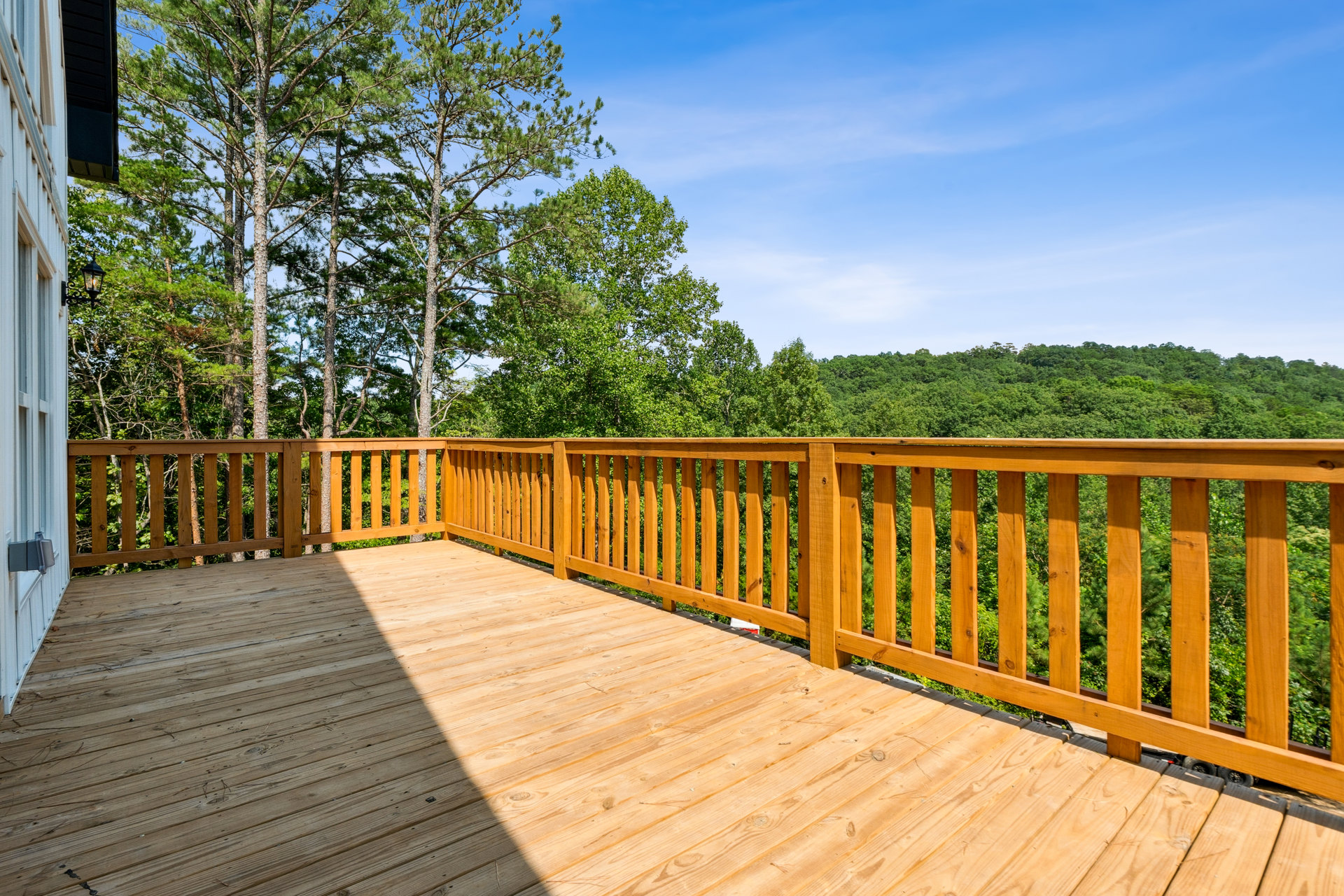 Wooden deck with railing overlooking a group of trees under a blue sky with clouds, white door with grey box, window facing tree-filled landscape