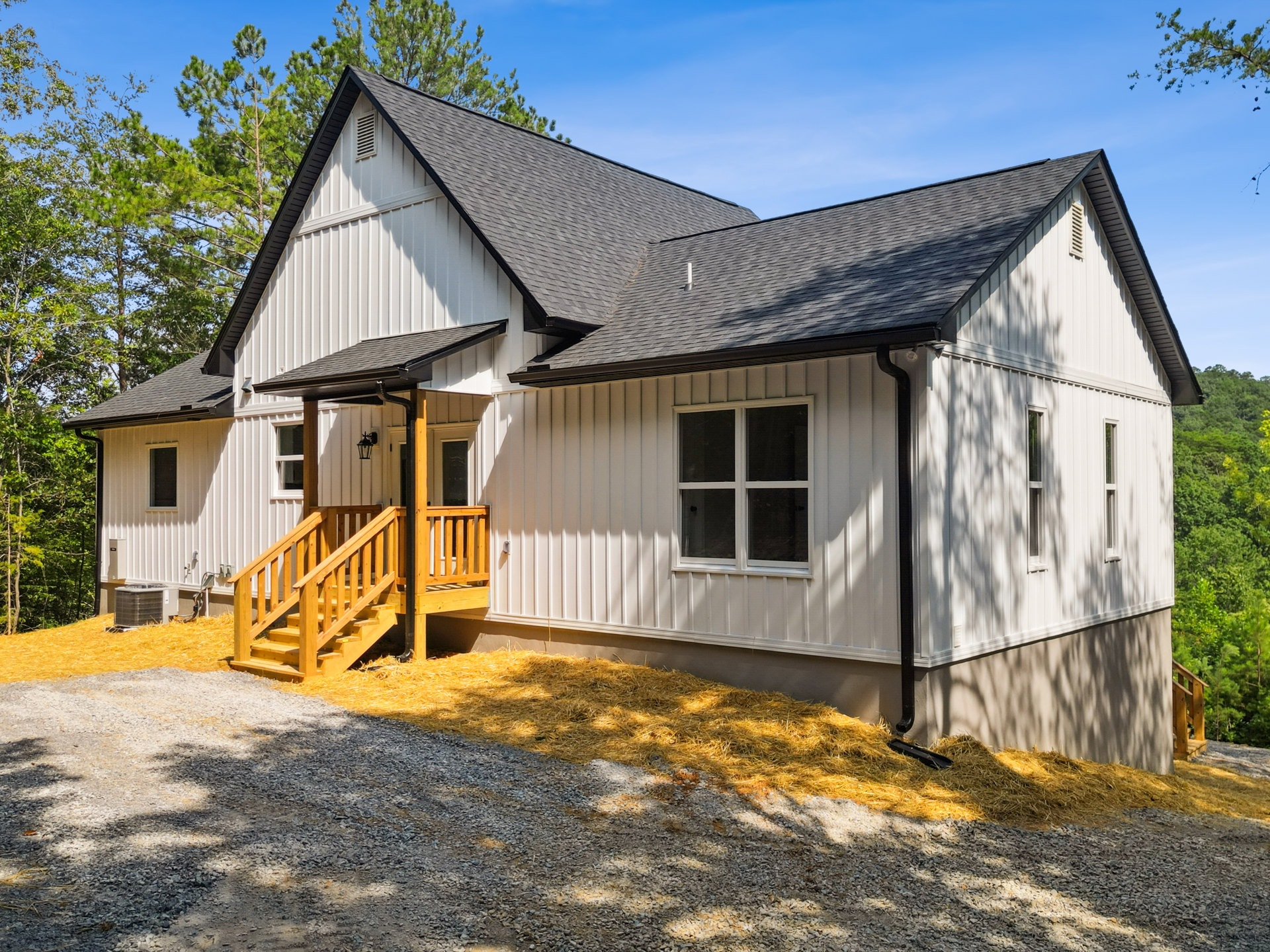 Two-story home surrounded by mature trees, wood exterior staircase leading to upper level, white-framed window, outdoor air conditioner unit, pile of hay on ground.