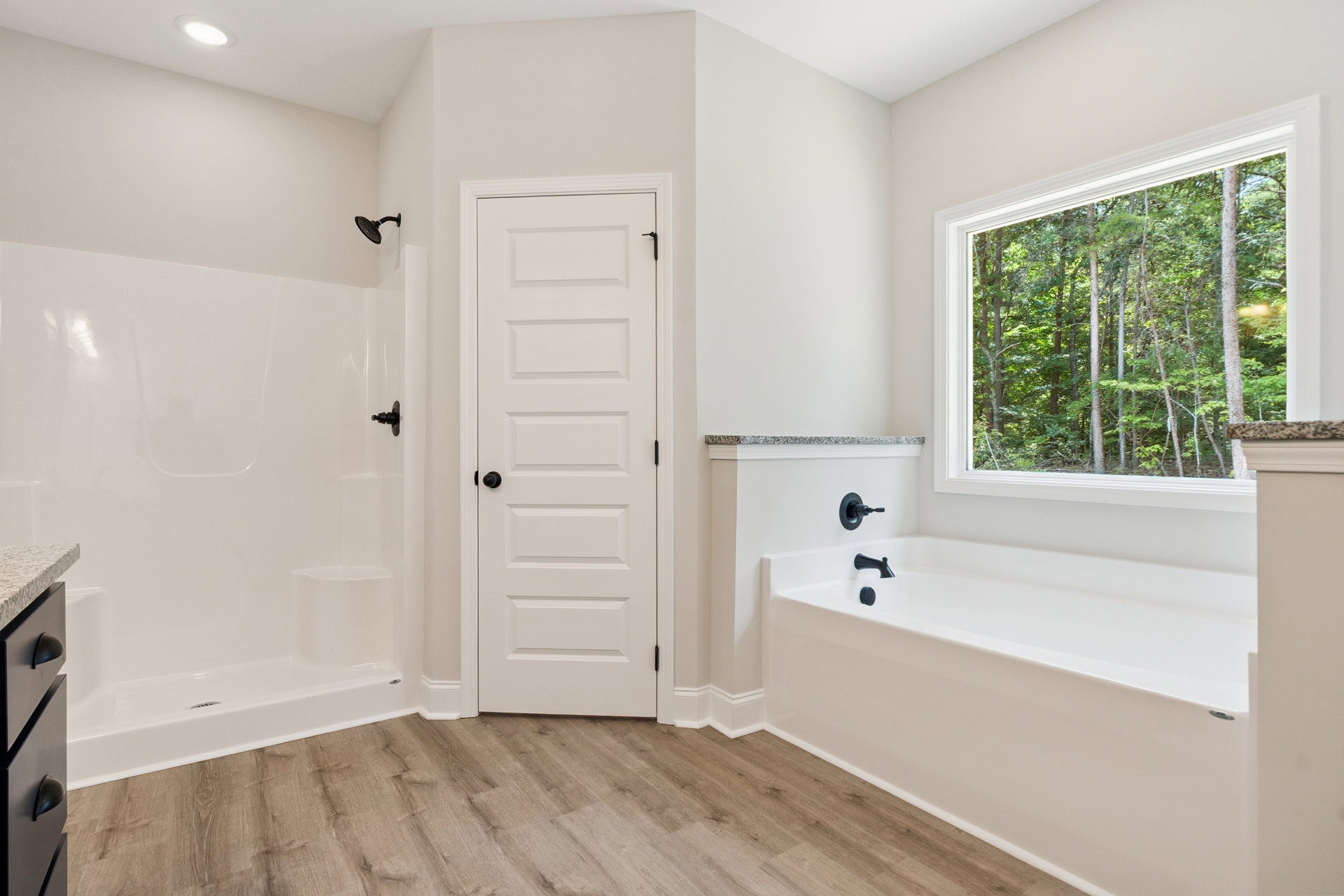 Bathroom with freestanding white bathtub and matte black faucet, glass-enclosed shower with black showerhead, white paneled door with black knob, window overlooking trees, light