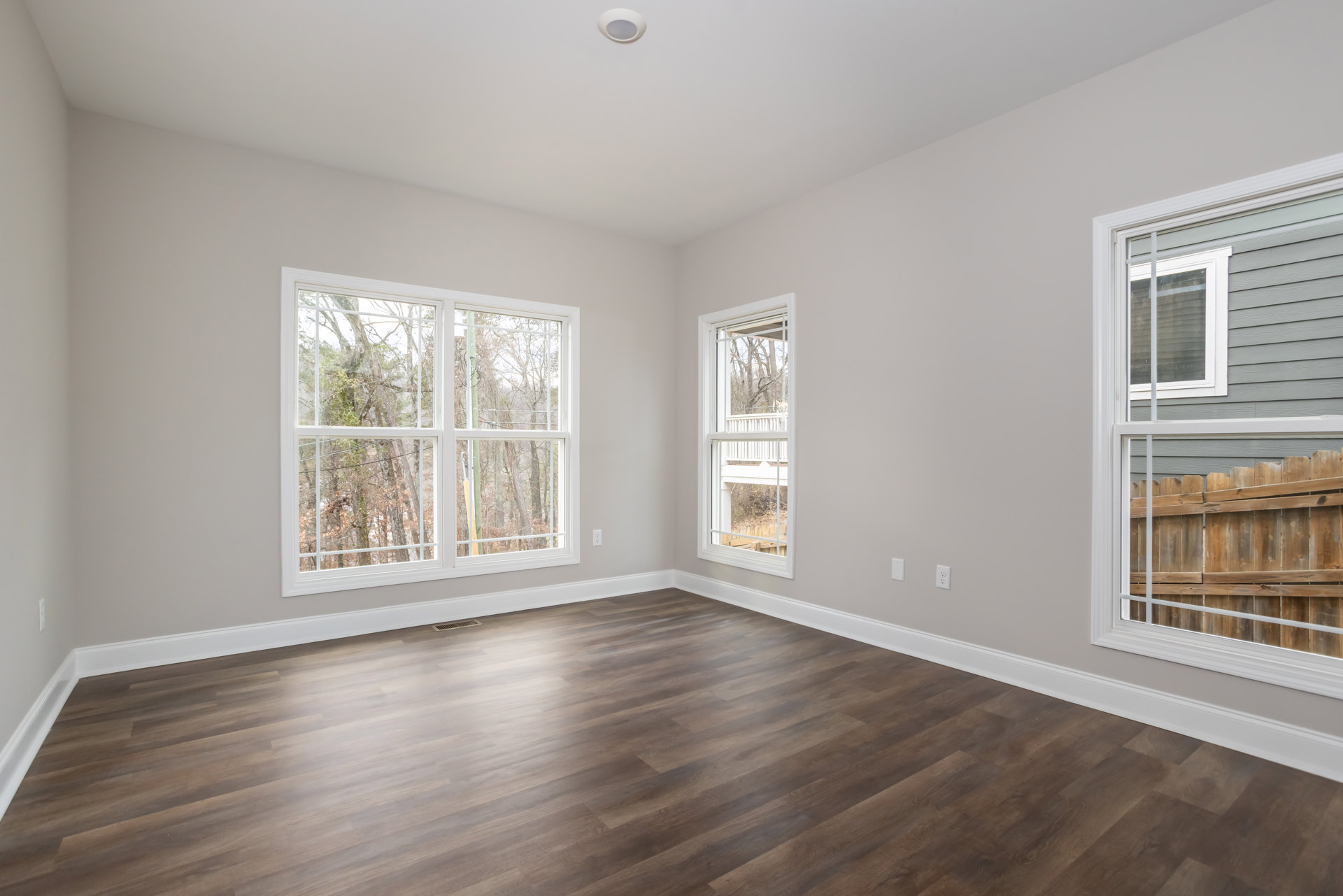 White walls and wood flooring in a room with a large window overlooking trees and a wooden fence, white railing visible outside.