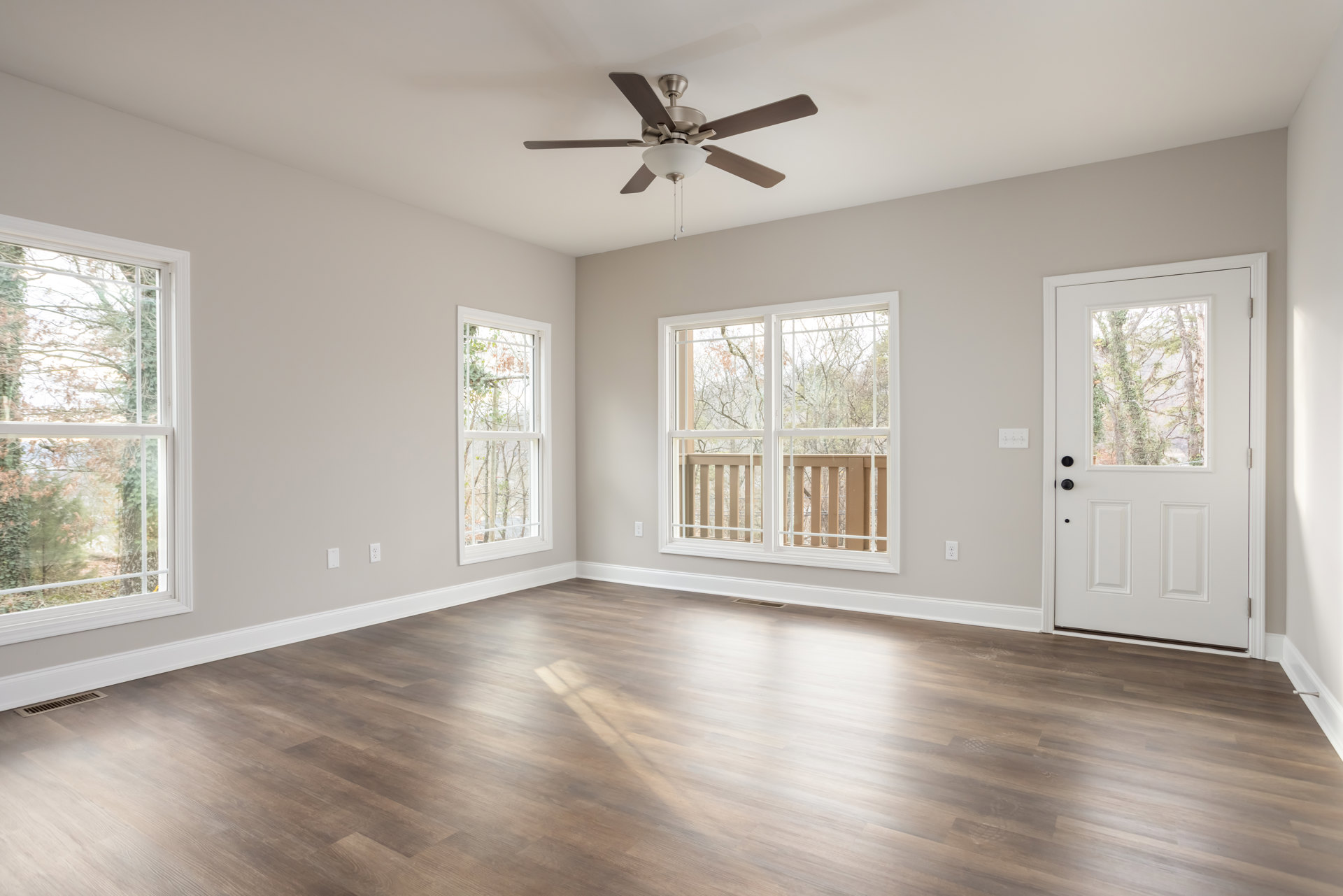 Ceiling fan with light fixture above wood flooring, large windows showing deck and trees, white door with glass panel, neutral walls and molding