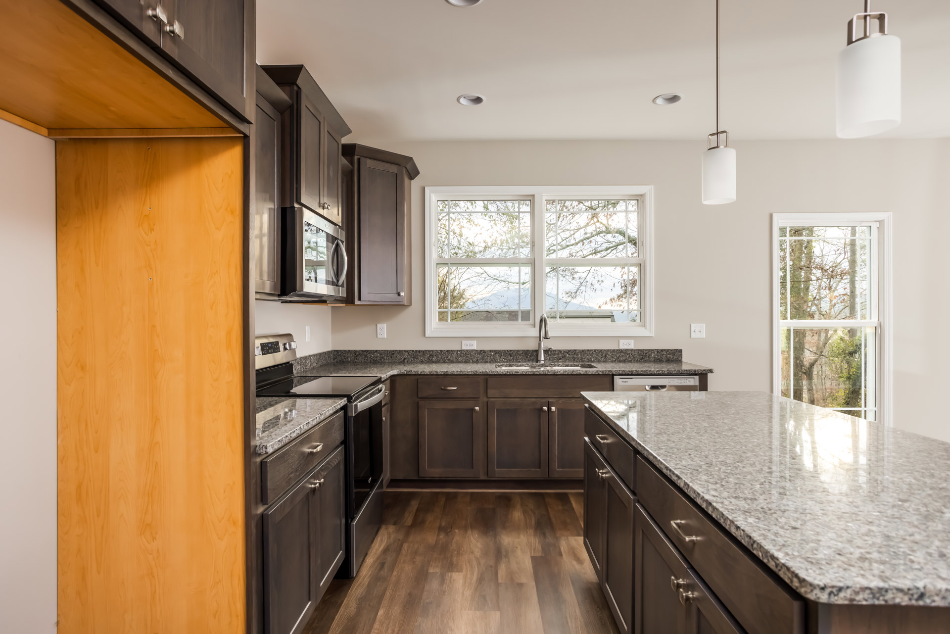 Granite countertops with black speckles, stainless steel sink beneath a window overlooking trees, wood flooring, white cabinetry, and modern kitchen appliances