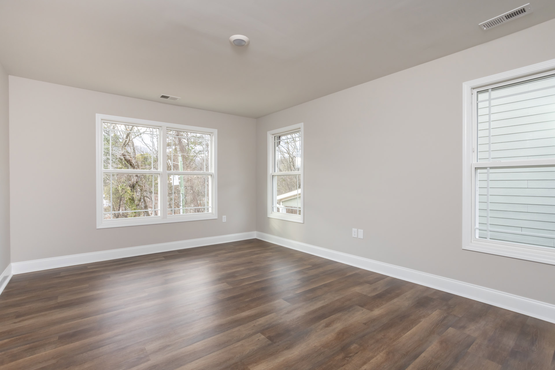 Wood flooring with white trim, large windows with blinds, sunlight streaming across the floor, ceiling vent, trees visible outside