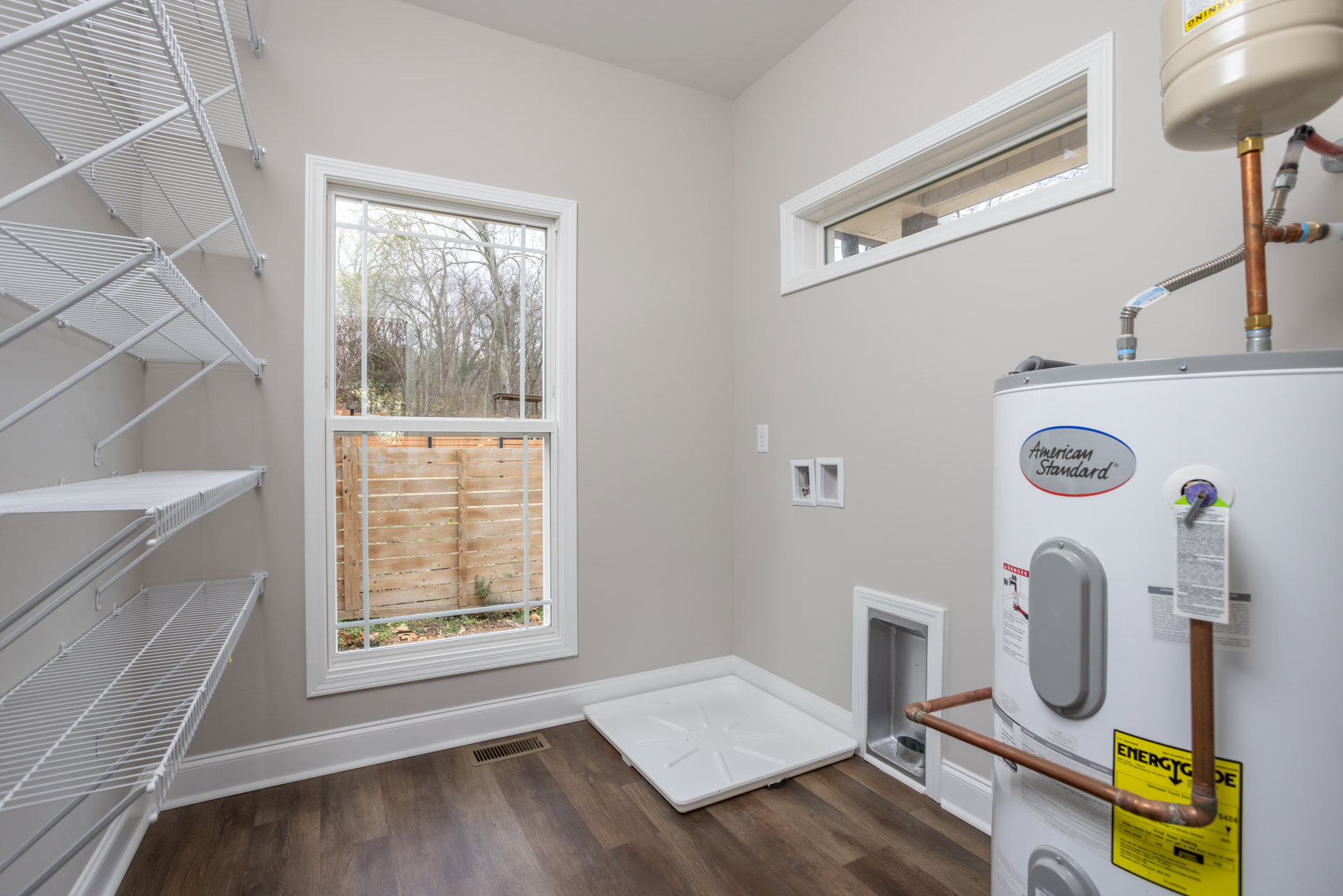 Laundry room with white washing machine, water heater tank, white plastic container on wood countertop, plaster walls, window overlooking wood fence and trees