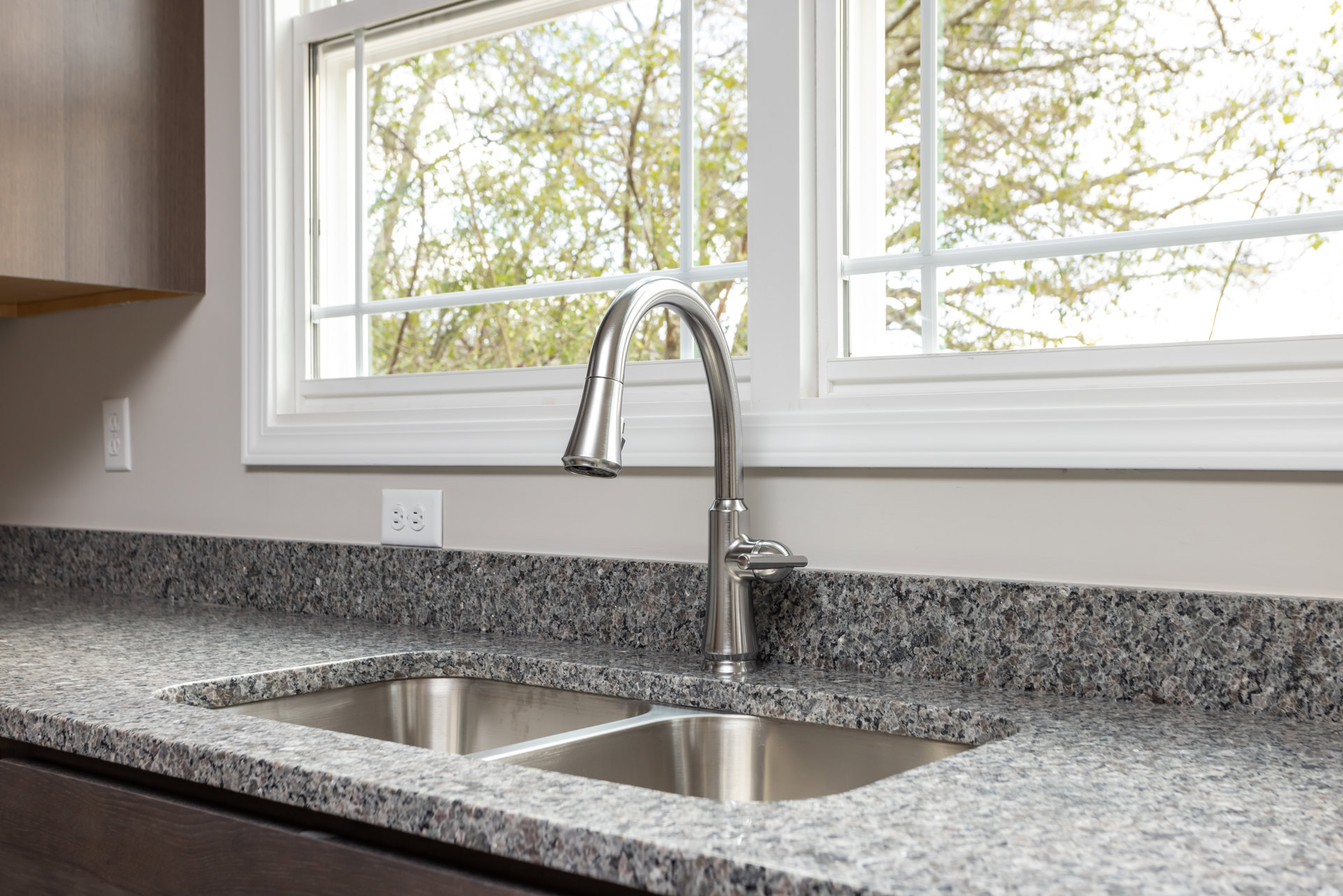 Stainless steel sink with modern chrome faucet set in quartz countertop, positioned beneath a large window with white tile backsplash and electrical outlet visible on wall