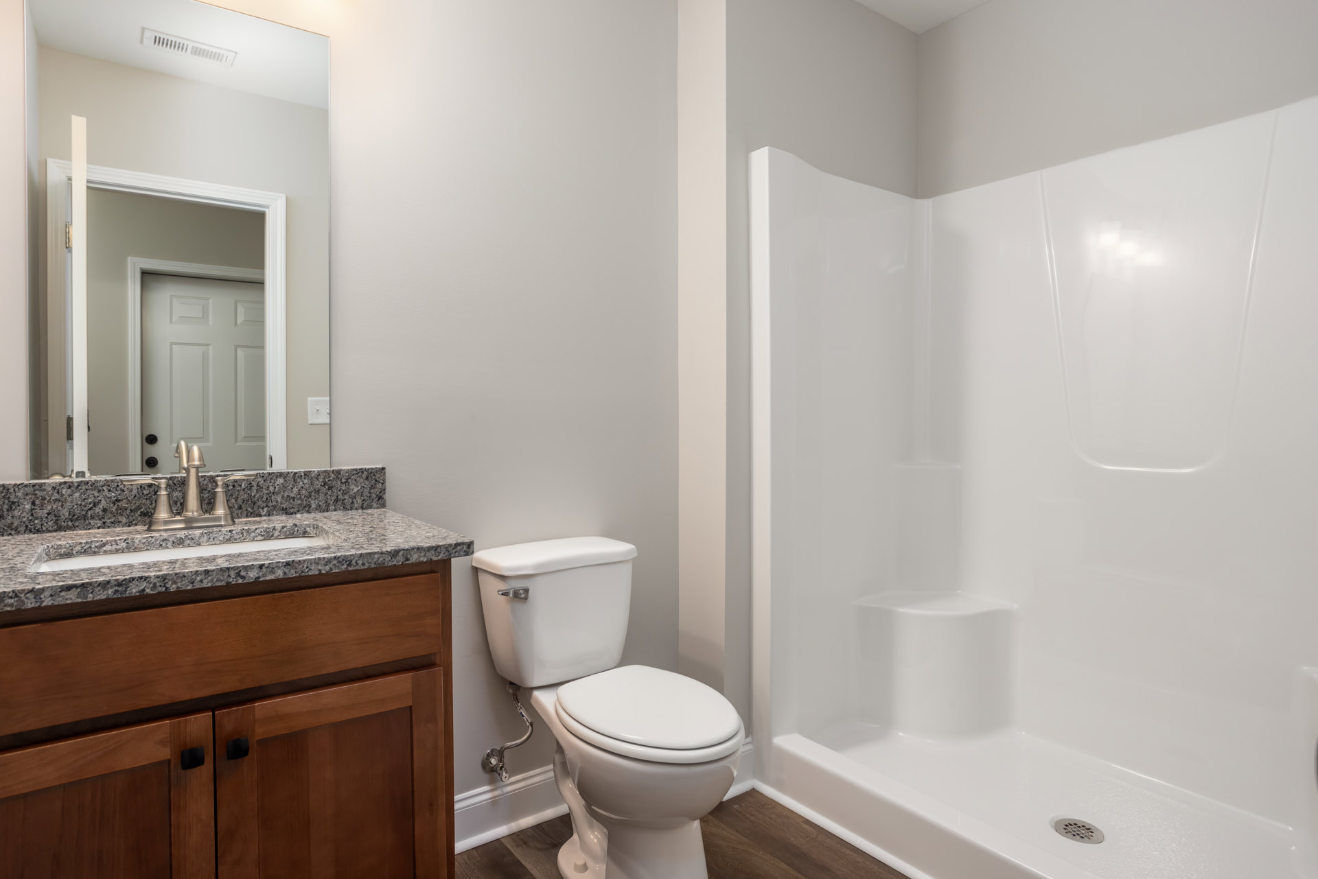 White toilet with lid up beside glass-enclosed shower, light tile flooring, wall-mounted sink with chrome faucet, large mirror above sink, neutral wall tiles
