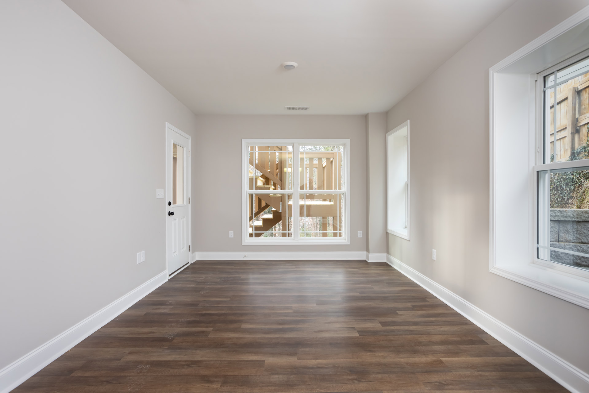 Wood flooring and white walls in a bright room with a large window, white door, and staircase visible in the background; outside the window, a fence, grass, and plants are seen.