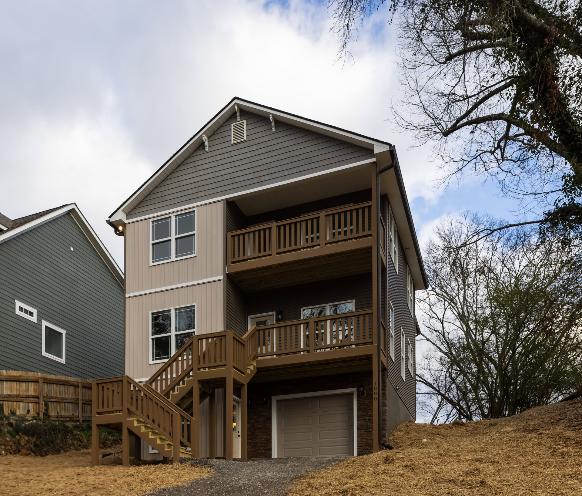 Brown wooden stairs lead to a porch on a custom home with a white-trimmed window, attached garage, and light siding, surrounded by trees under a partly cloudy sky.