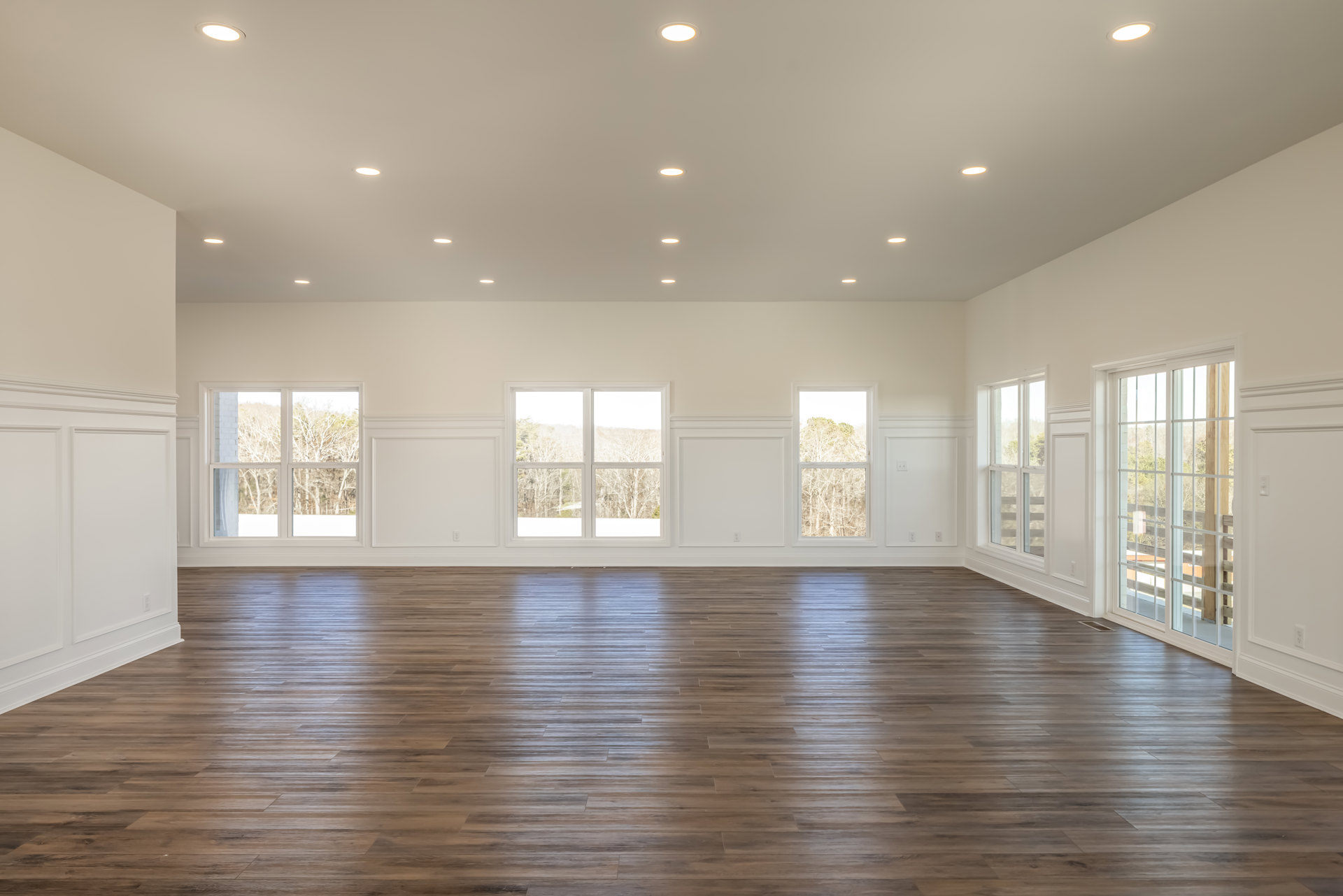 Spacious room with white plaster walls, hardwood flooring, large window overlooking trees, and a row of recessed ceiling lights