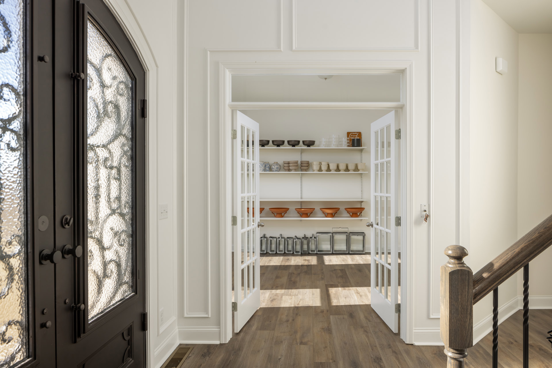 White walls and built-in white shelves displaying bowls and utensils, wood flooring and railing, nearby door and window with metal hardware