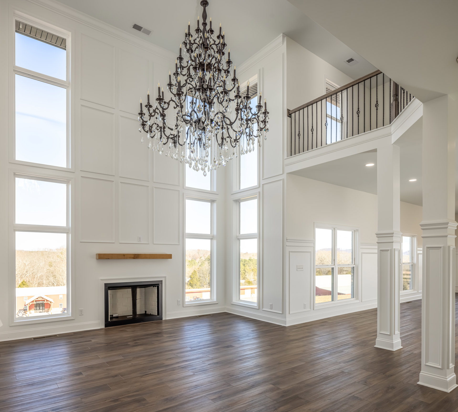 Spacious living room with wood flooring, ornate chandelier hanging from ceiling, white fireplace with metal pipe, large windows overlooking trees, decorative wall molding