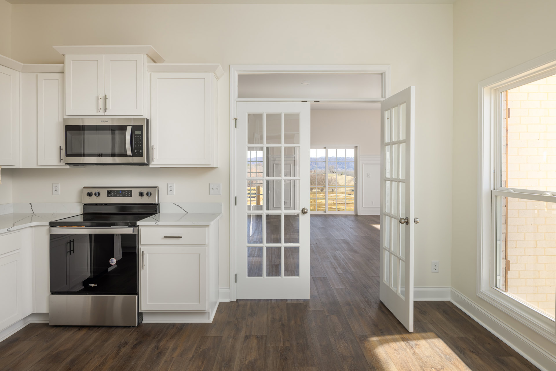 White kitchen cabinets, open microwave door, black and silver oven, window with screen, white door with glass panes and silver handle, light countertops and flooring