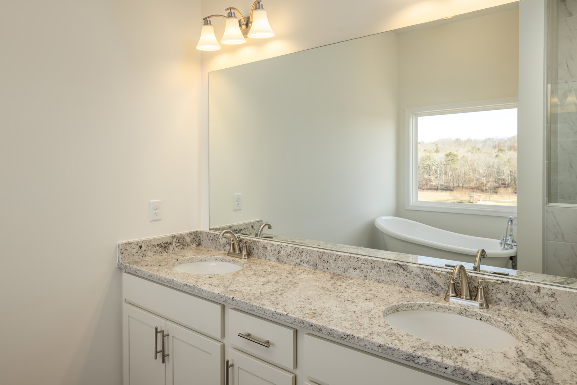 Bathroom with double sinks set in a stone countertop, large wall mirror above, three-light fixture, white bathtub, window overlooking trees, tile flooring, and wood cabinetry.