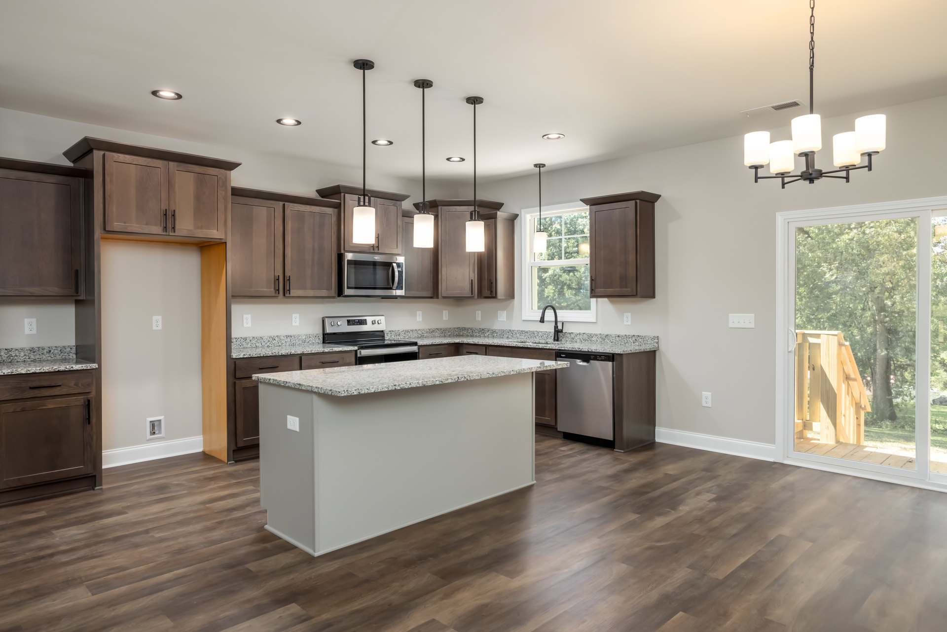 Spacious kitchen featuring a large central island with built-in stove and oven, wood flooring, white cabinetry, stainless steel microwave, and a window overlooking trees with wood