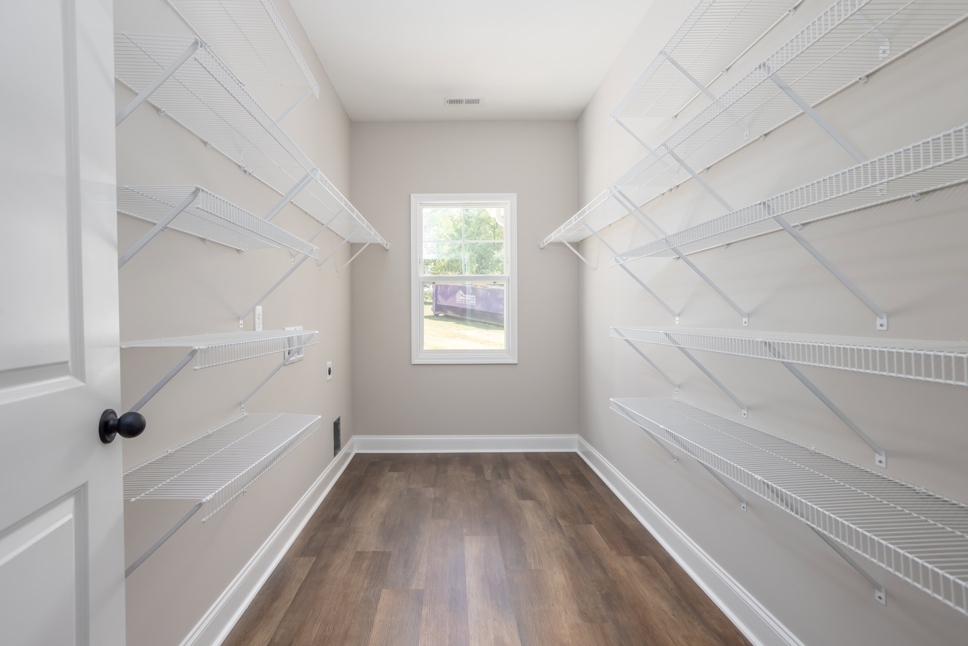 White-walled room with built-in white shelves, wood flooring, large window overlooking construction equipment and a container outside