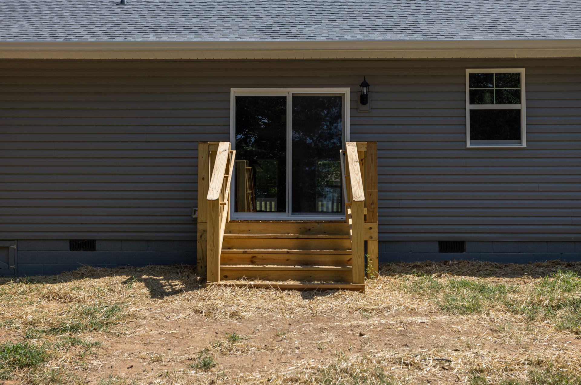 Wooden stairs with natural finish leading to a house entrance featuring a glass-paneled door, white-framed window, and horizontal siding; grassy yard and plants surround the porch