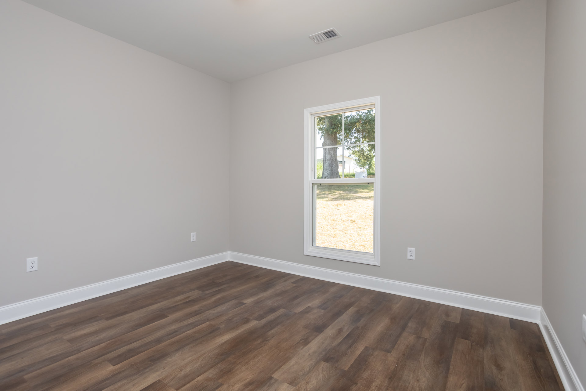 Sunlit room featuring a large window overlooking a grassy yard with a tree and white fence, hardwood flooring with white baseboard trim, and a ceiling vent.