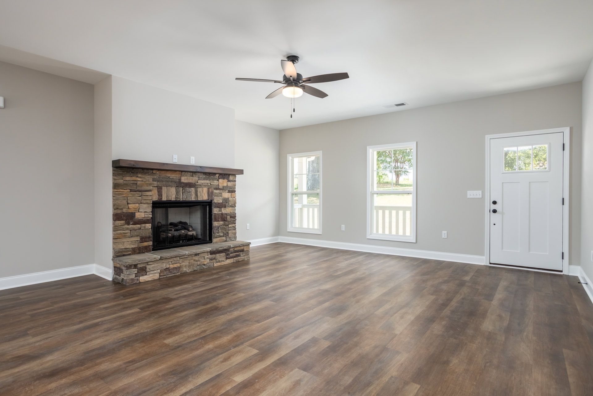 Living room with wood flooring, stone fireplace filled with logs, stone staircase, ceiling fan with light, white door with window, large window showing tree outside