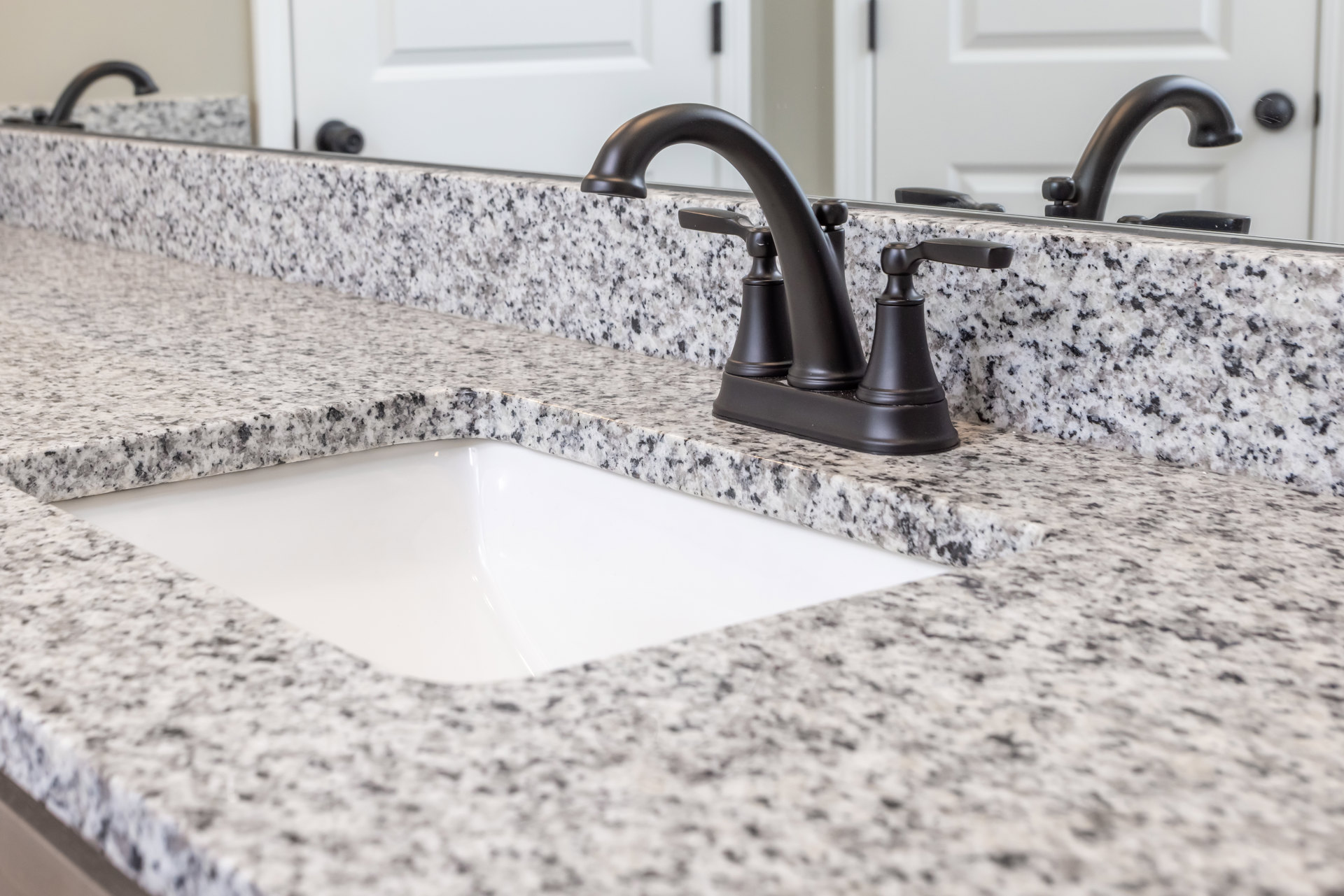 Black faucet and rectangular white sink set in a granite countertop, with light gray tile backsplash and wall-mounted plumbing fixtures in a modern bathroom.