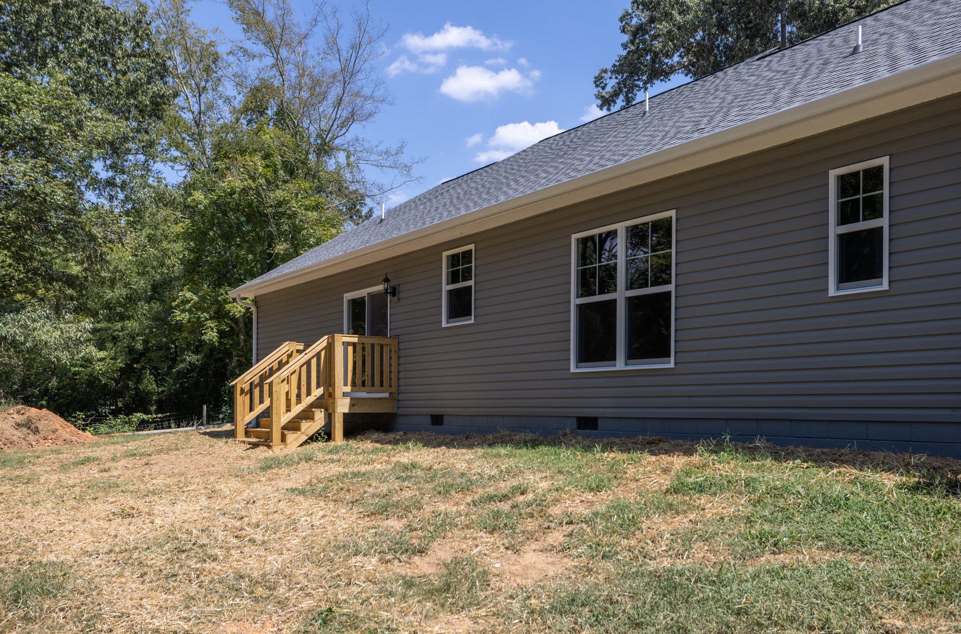 Wooden staircase leading to a covered porch, white-framed windows reflecting trees, horizontal siding exterior, grassy yard with patches of dirt