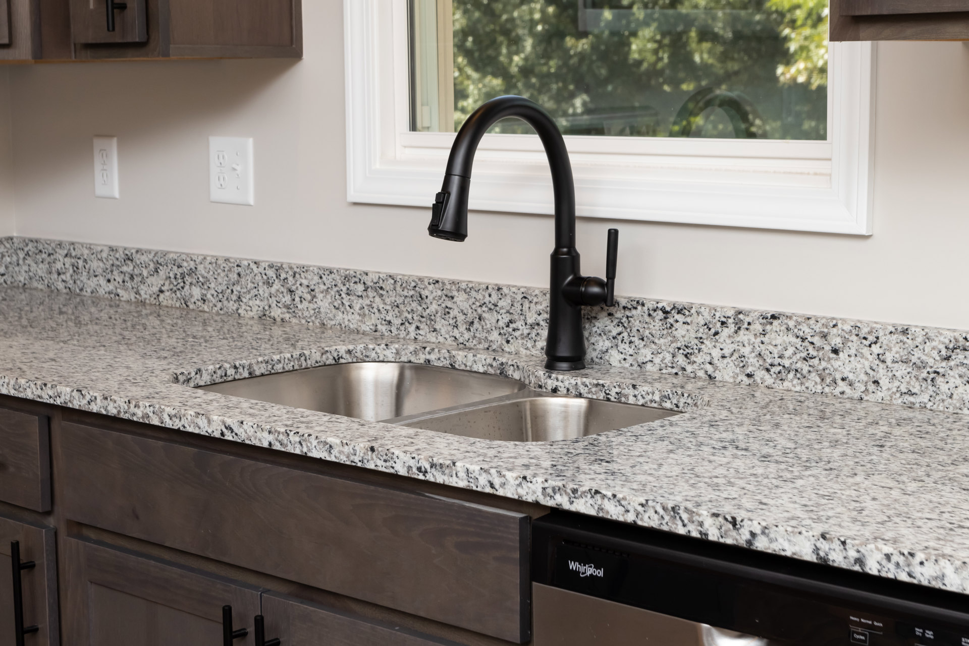 Black faucet and stainless steel sink set in speckled granite countertop, white electrical outlet on backsplash, window with view of green trees, white cabinetry below.