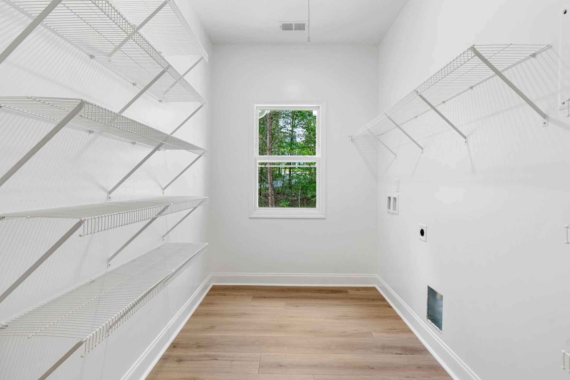 White built-in shelves with metal supports line a wall in a room featuring light wood flooring, white plaster walls, and a large window overlooking leafy trees.