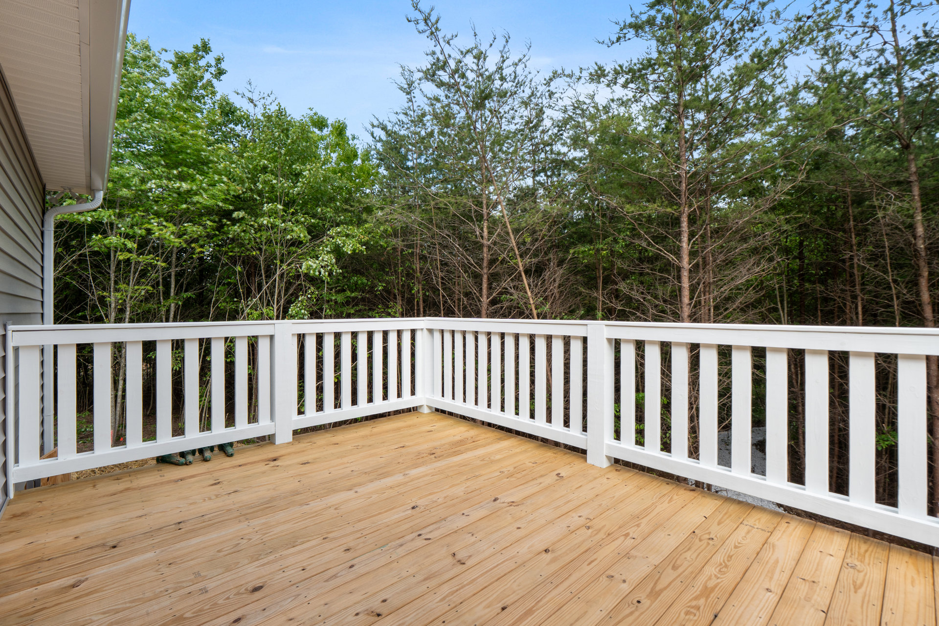 Wooden deck with white railing overlooking a group of trees, blue sky visible above, exterior of house partially shown