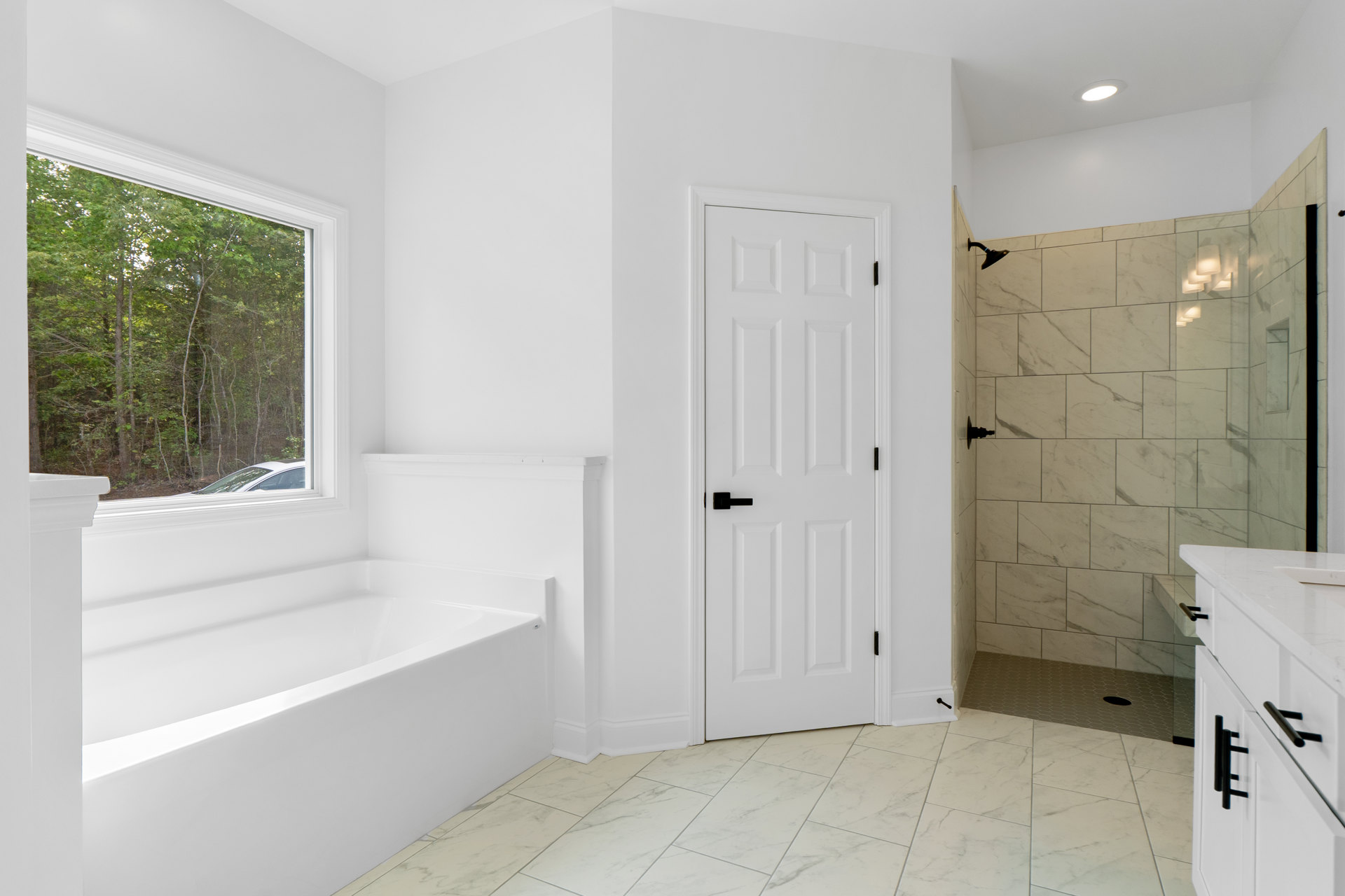 Bathroom with white tile floor, freestanding white bathtub beneath a window showing trees, white cabinetry, and a white door with black handle