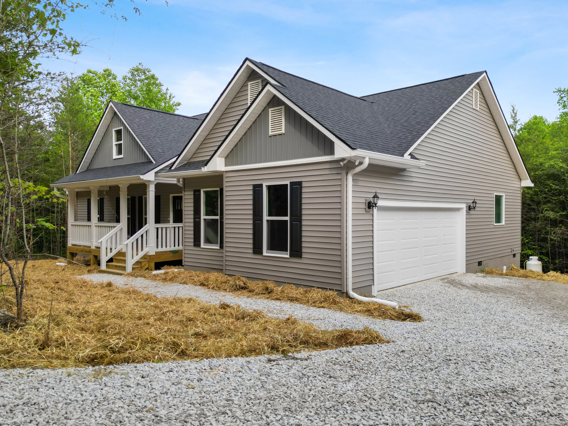 Two-story house with white garage door, paved driveway, gray shingle roof, white-framed windows, outdoor lamp beside garage, gravel path bordered by grass, trees and Robert Frost