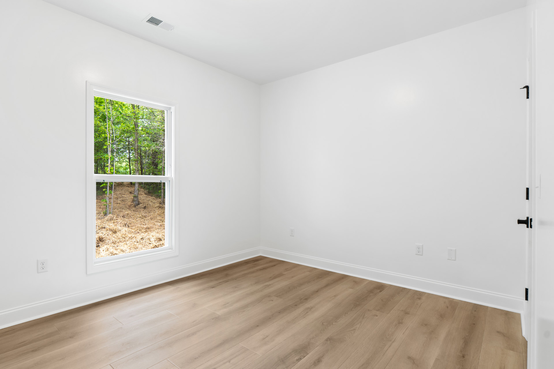 White-walled room with wood laminate flooring, large window overlooking trees and grassy landscape, white ceiling