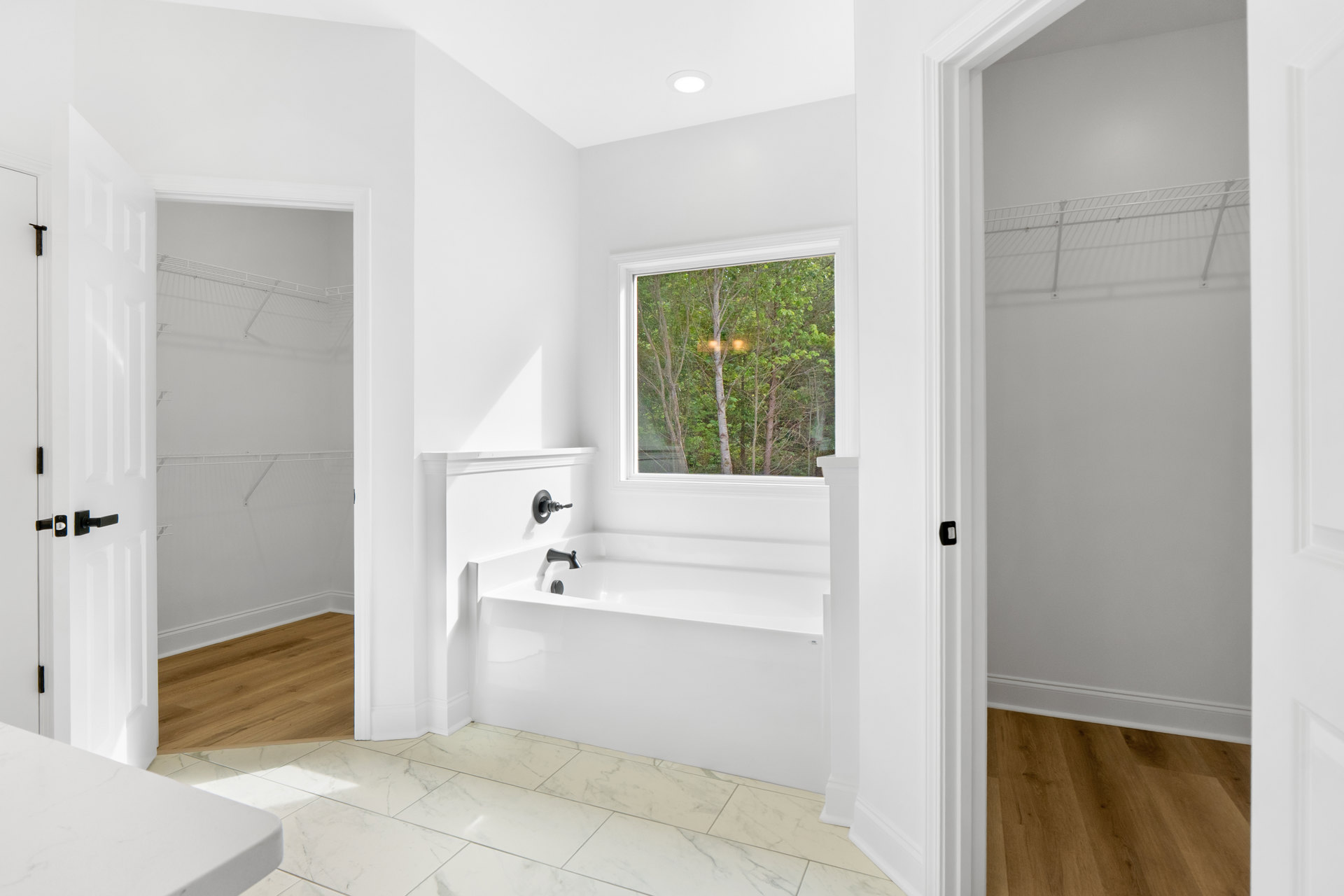 Bathroom featuring a freestanding bathtub and glass-enclosed shower, large window with tree views, wood flooring with white trim, and a white side table near a wall switch.