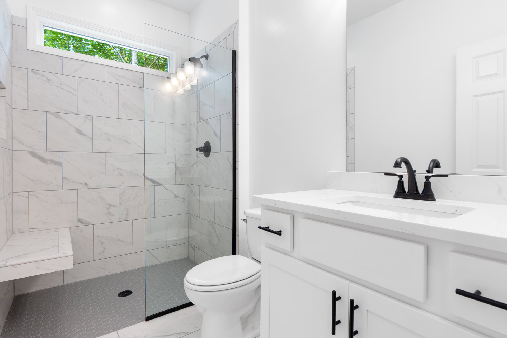 Bathroom with glass shower enclosure, white toilet, white sink with black handles, tile flooring, window showing trees outside