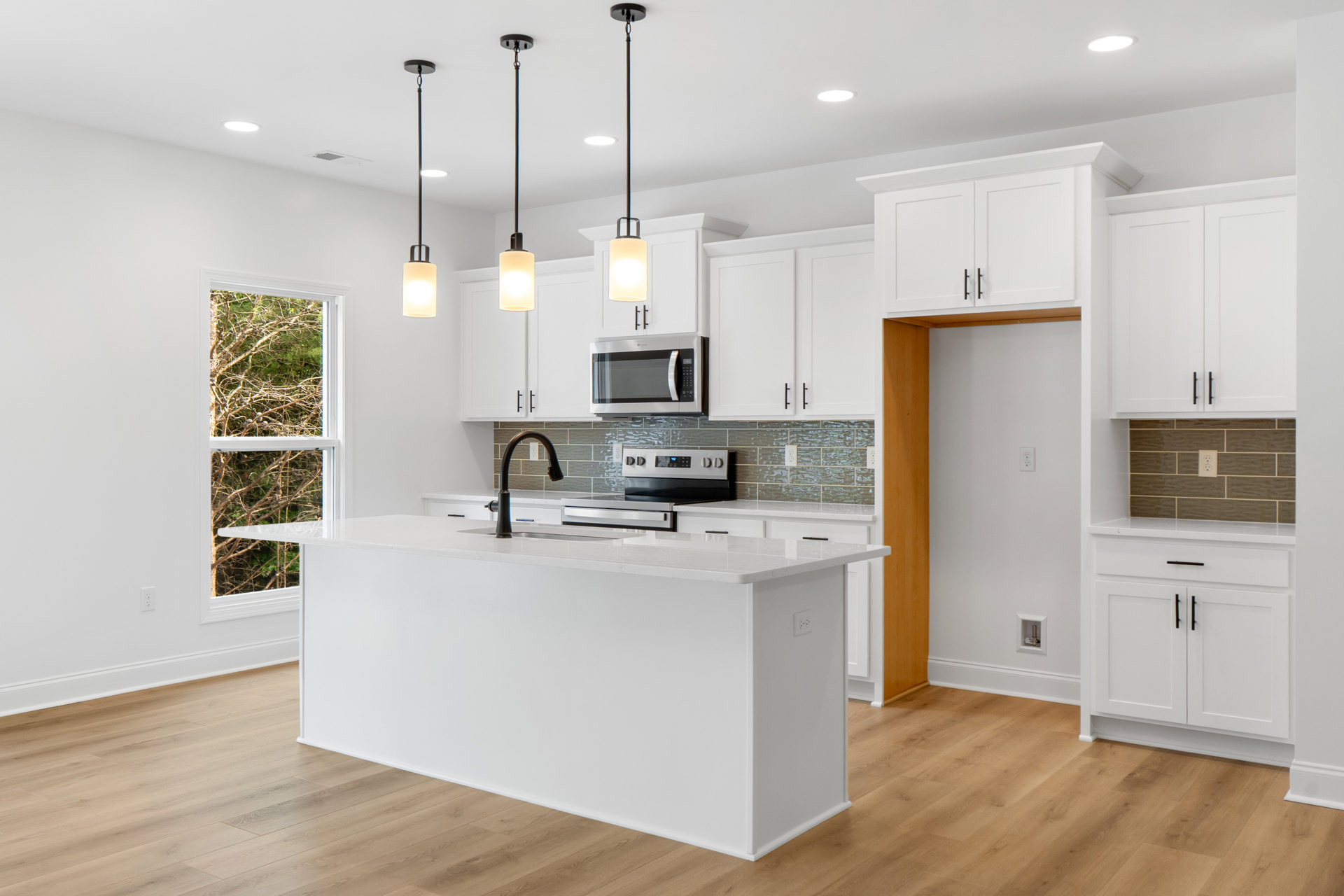 White kitchen with shaker cabinets, wood plank flooring, tile backsplash, black countertop lamp, built-in microwave, stainless sink, and illuminated pendant light.