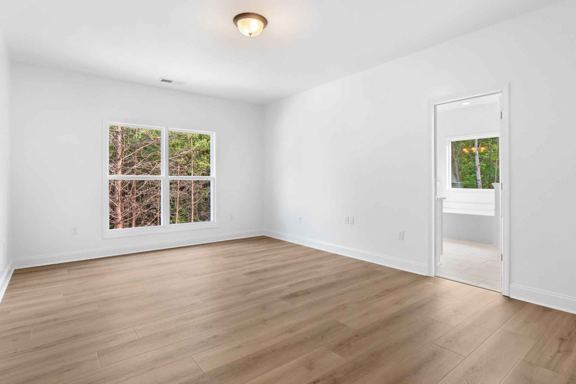 Room with hardwood floor, large window overlooking trees, round ceiling light fixture, white plaster walls, and door with glass panel.