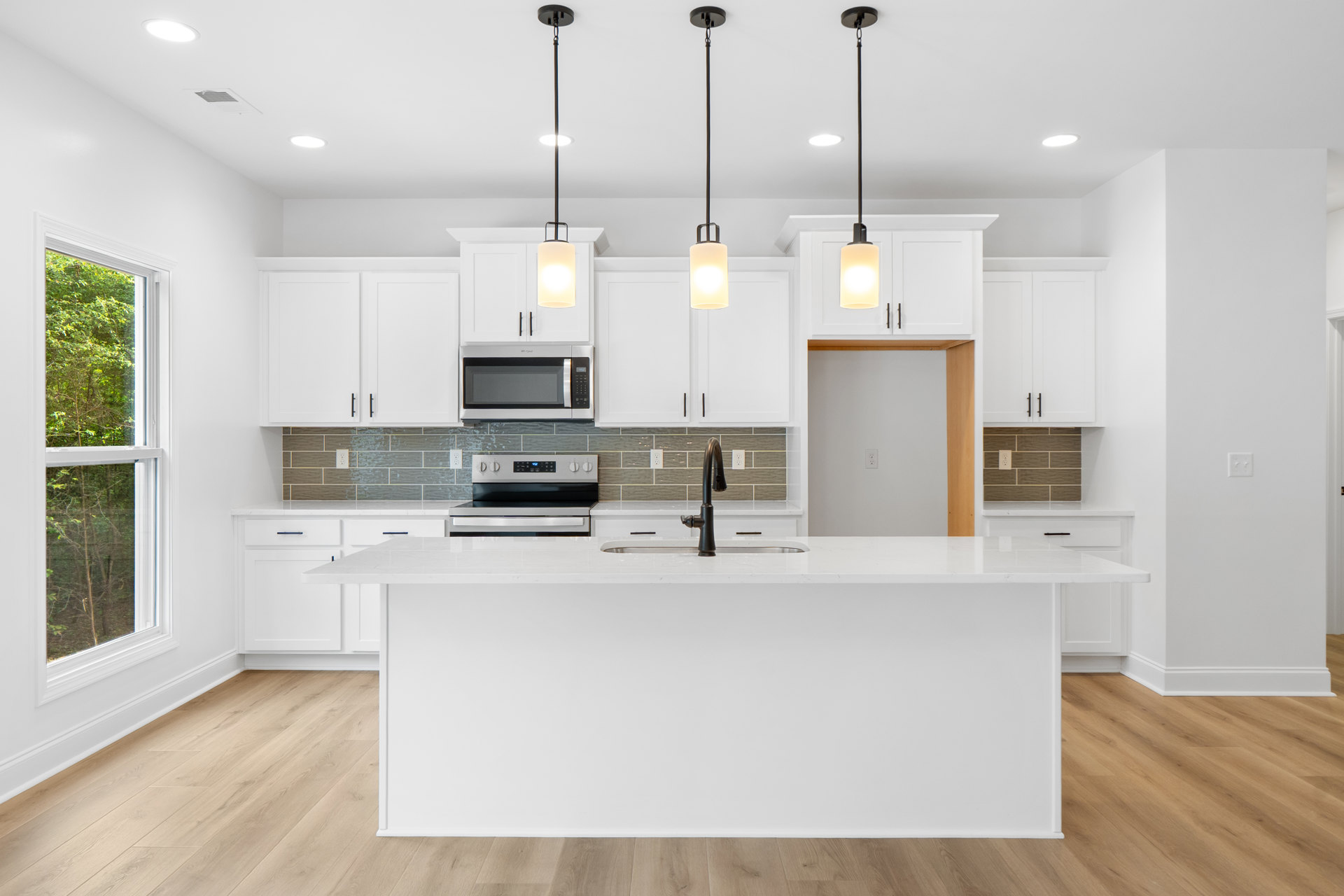 White kitchen with shaker cabinets, quartz island, stainless steel oven and microwave, chrome faucet, circular pendant light, light hardwood flooring.