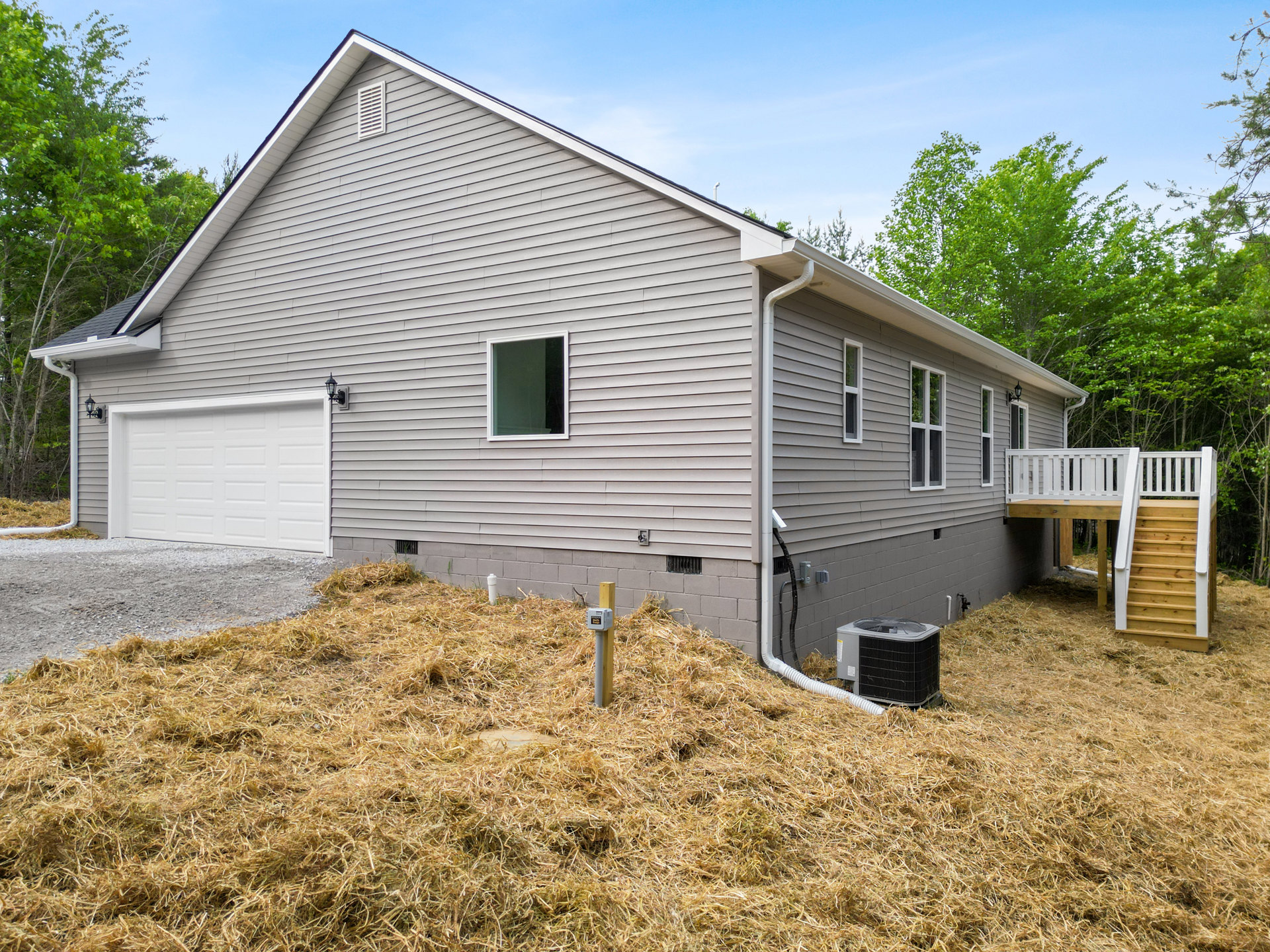 Two-story house with light siding, wooden deck featuring white railing, white-framed windows, white garage door, fenced yard, air conditioner unit on ground, pile of hay beside