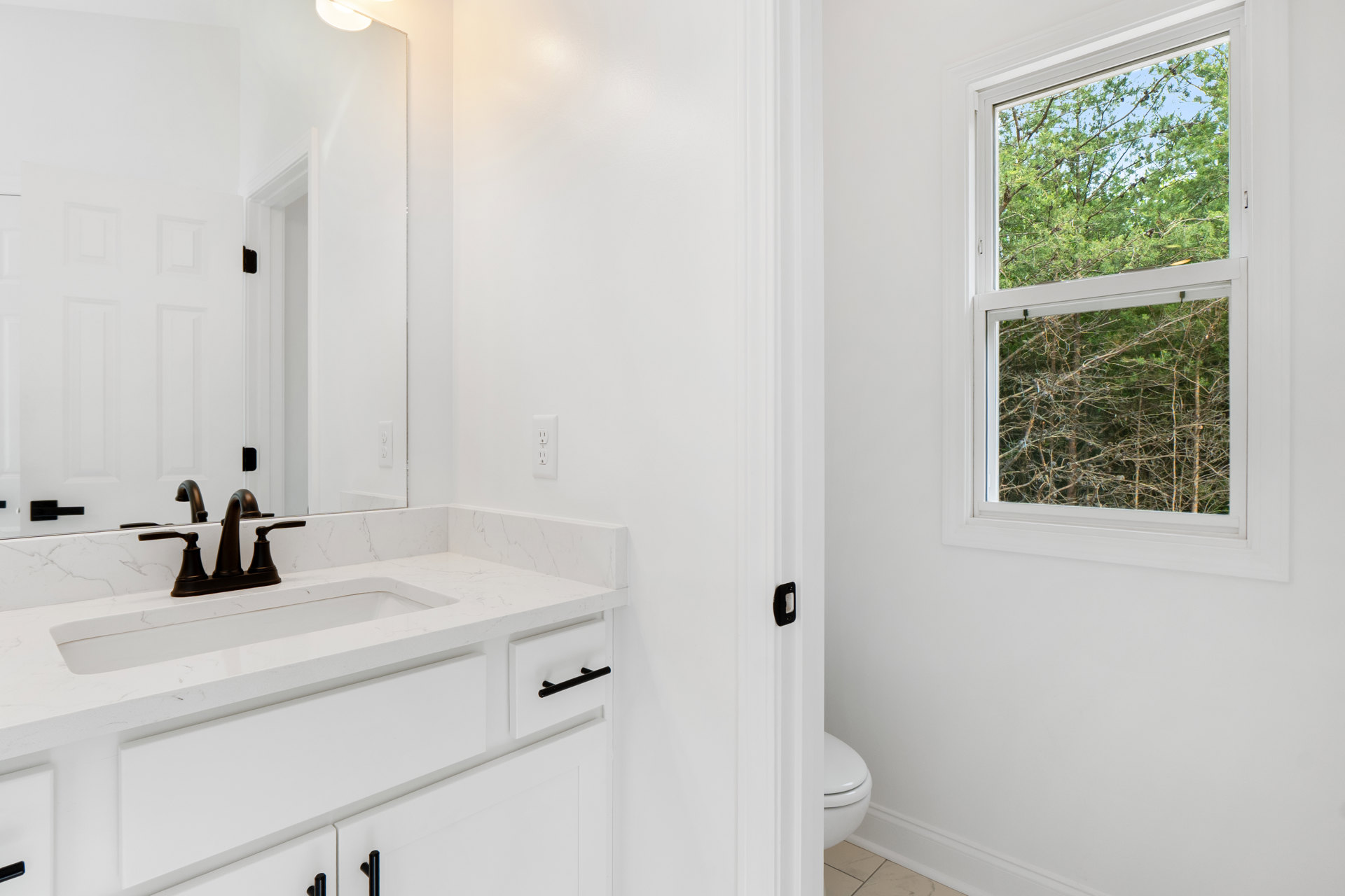 White countertop with black faucet and rectangular mirror above, toilet beside sink, window overlooking green trees, light tile flooring