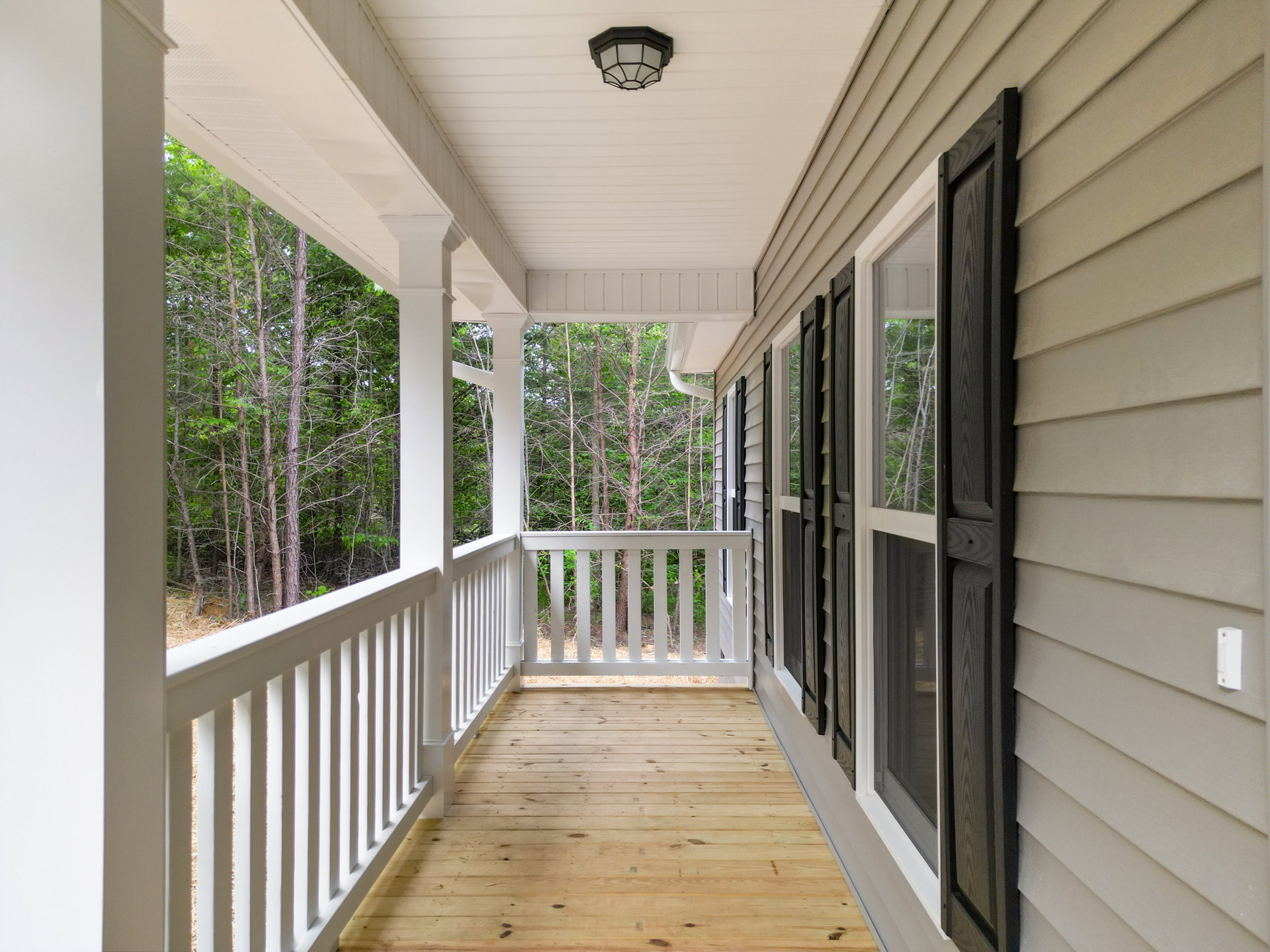 Wooden porch deck with white railing, black light fixture mounted on post, row of windows, leafy trees visible in background