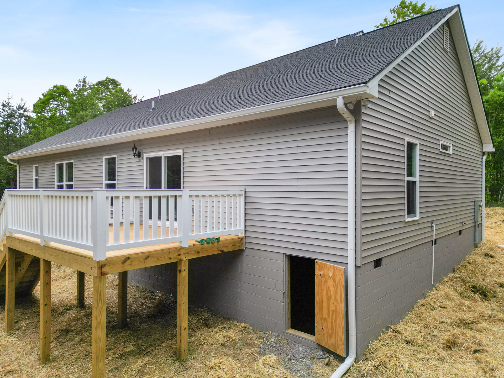 Two-story house with gray siding, white-trimmed windows, wooden front door with black screen, elevated deck featuring white railing, green glass window, and gabled roof surrounded