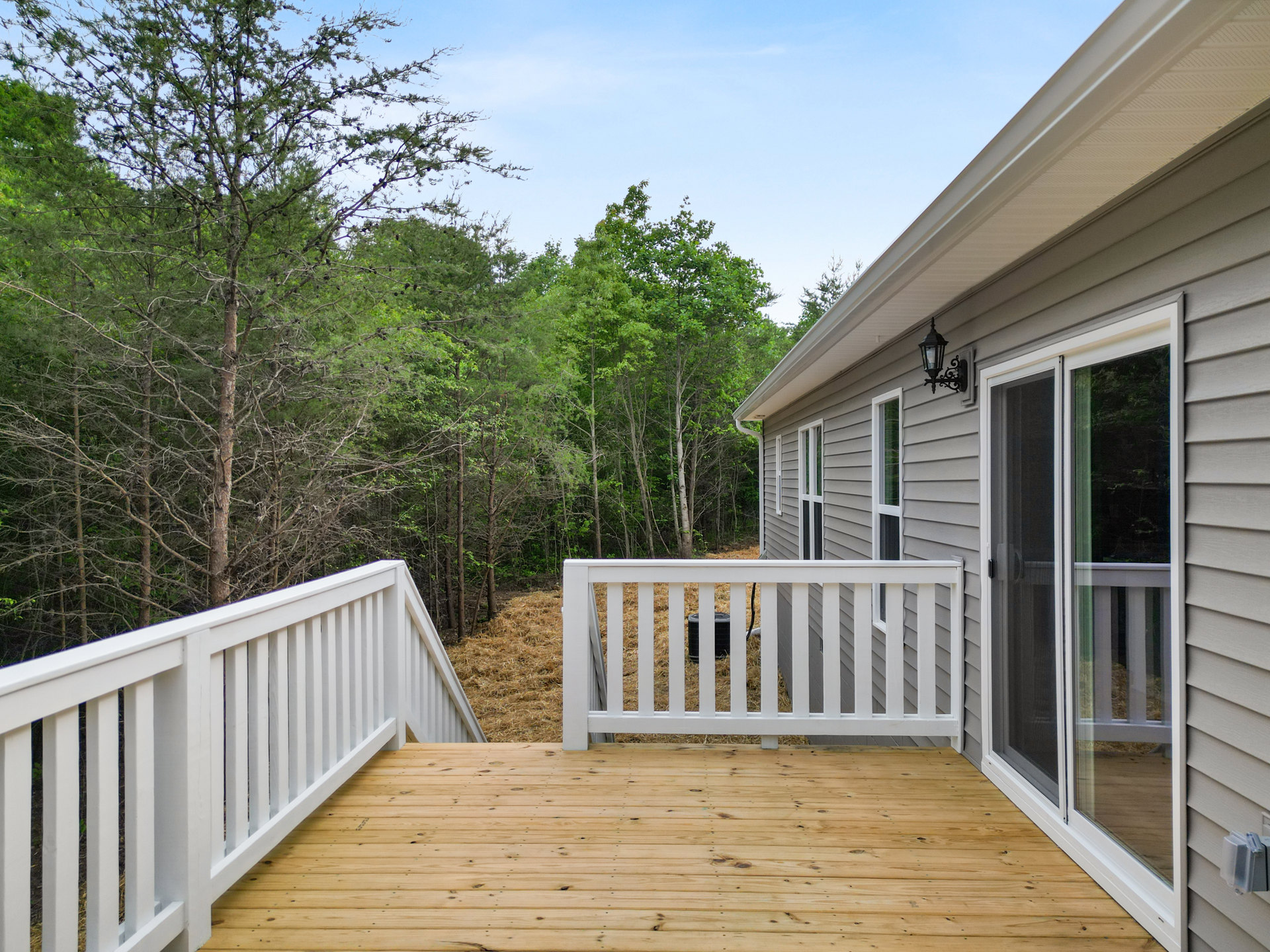 Wooden deck with white railing, sliding glass door, lamp post, black barrel on grass, light-colored siding, mature trees in background