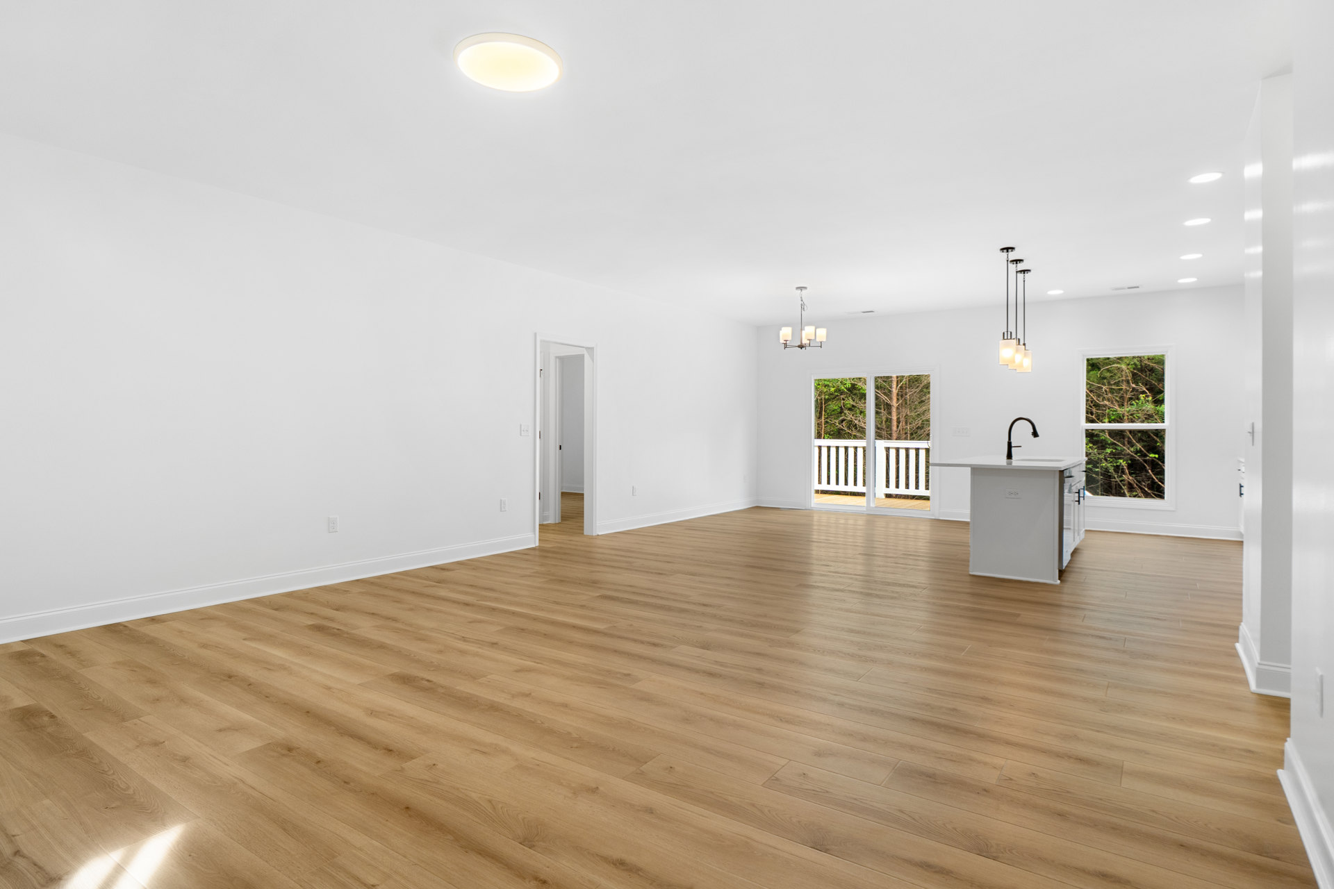 Spacious room with light hardwood floors, white walls, large windows overlooking trees, and a white kitchen island with a black faucet